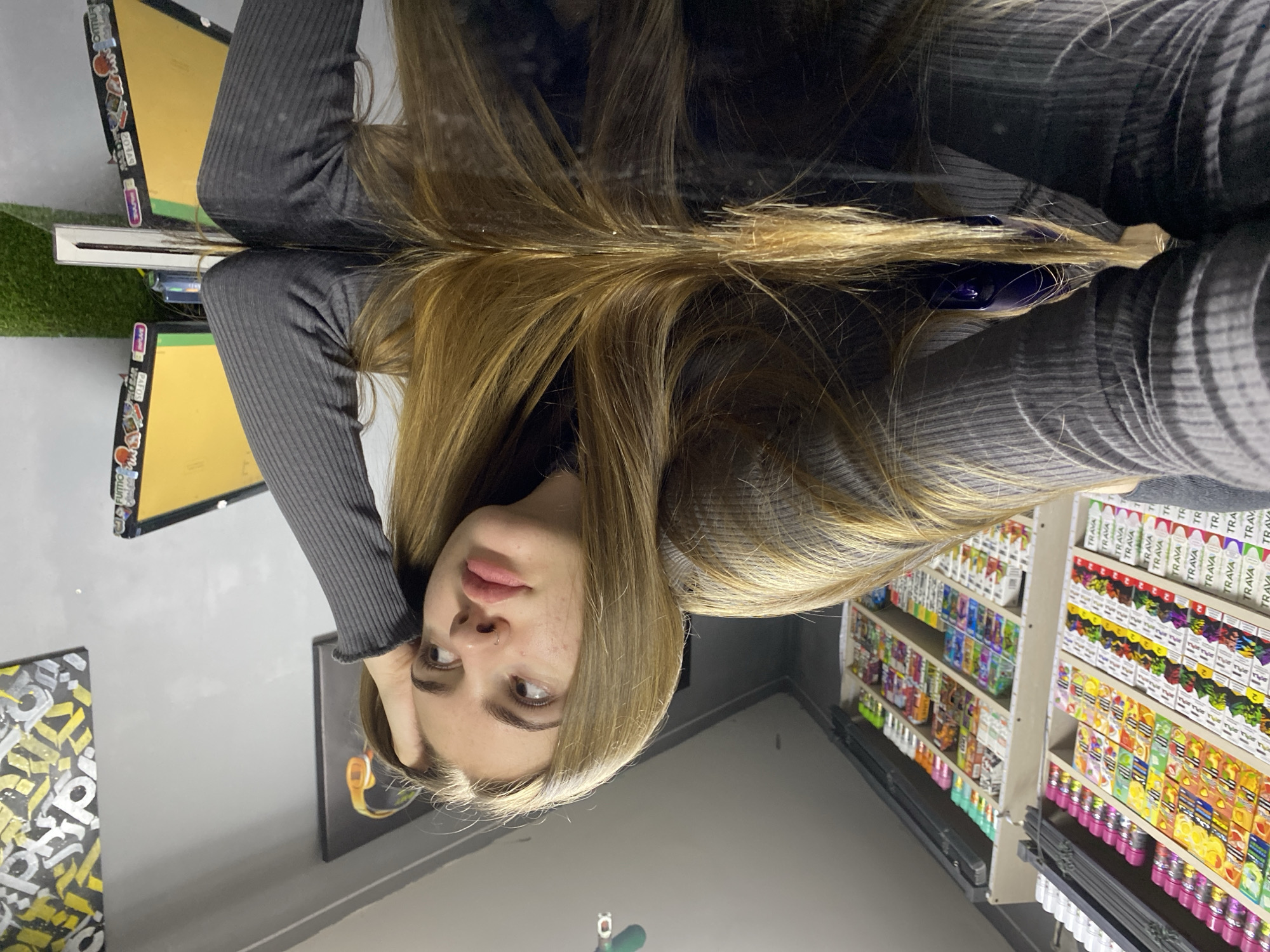 The image shows a young woman with long, blonde hair lying on a reflective black surface, likely a table. She is wearing a grey ribbed long-sleeved shirt and has her hand resting on her head. Her gaze is directed away from the camera, with a slightly contemplative or bored expression. The background reveals shelves filled with numerous colorful products, possibly vape juices or similar retail items. To the right, a computer monitor is visible, adorned with stickers. The environment appears to be an indoor retail space, potentially a vape shop or convenience store, given the product display. The location is identified as Dyakovo, Russia.