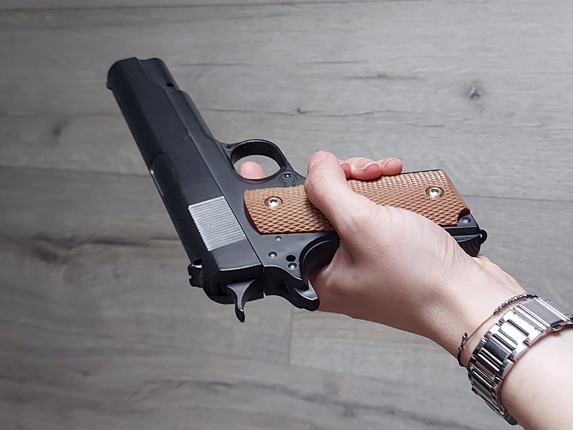 A black pistol with brown grips is being held in a person's hand. The person is wearing a silver watch and a bracelet. The background is a gray surface that appears to be wood-grain flooring. There is no visible evidence to suggest the location is Izhevsk, Russia.