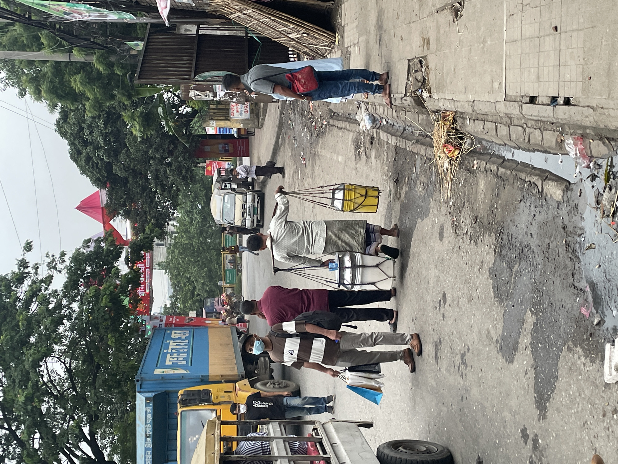 The image shows a street scene in Dhaka, Bangladesh. Several people are walking along a road with standing water and debris. A man carries a yellow container suspended by ropes. Another man carries a white drum. Several cars and trucks are in the road. Banners and trees are visible. A person wearing a face mask is walking along the road carrying shopping bags.