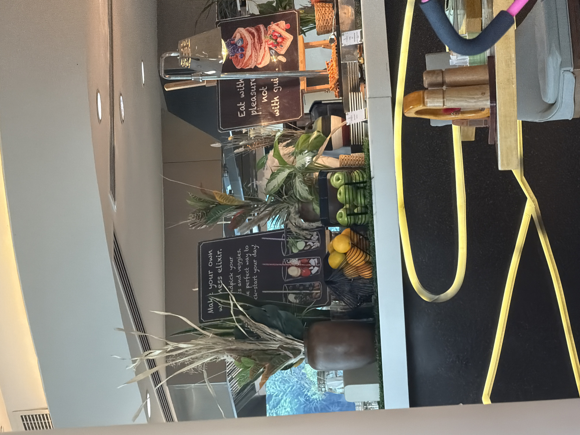 The image shows a buffet or self-serve food station in a restaurant, likely in Dubai, United Arab Emirates, based on the ambient lighting and modern aesthetic which is common in Dubai's hospitality sector. In the foreground, a yellow neon light fixture curves across the black countertop. Behind this, on the countertop, are two wooden salt and pepper shakers. To the right, a black handlebar with a pink grip is visible, suggesting part of a piece of equipment or furniture.
Further back, a black chalkboard sign with white text reads: "Make your own wellness elixir. Handpick your fruits and veggies. The perfect way to kick-start your day!" Below this sign, three illustrated glasses show layers of fruit and possibly other ingredients. To the left of the sign is a large brown ceramic vase filled with dried grasses and green leafy plants. In front of the vase, a black wire basket holds several orange fruits, possibly oranges or mandarins, and a smaller basket holds green apples.
To the right of the "wellness elixir" sign, another black chalkboard sign displays an illustration of a stack of pancakes topped with blueberries and a strawberry, and a waffle with strawberries and blueberries. The text on this sign reads: "Eat with pleasure not with gui". This suggests a dessert or breakfast offering. Behind this sign, a metal stand holds a silver lamp with a woven shade, and a small potted plant. The background shows kitchen equipment and a blurred reflection of greenery. The overall scene depicts a health-conscious or indulgent food presentation, common in upscale dining establishments.