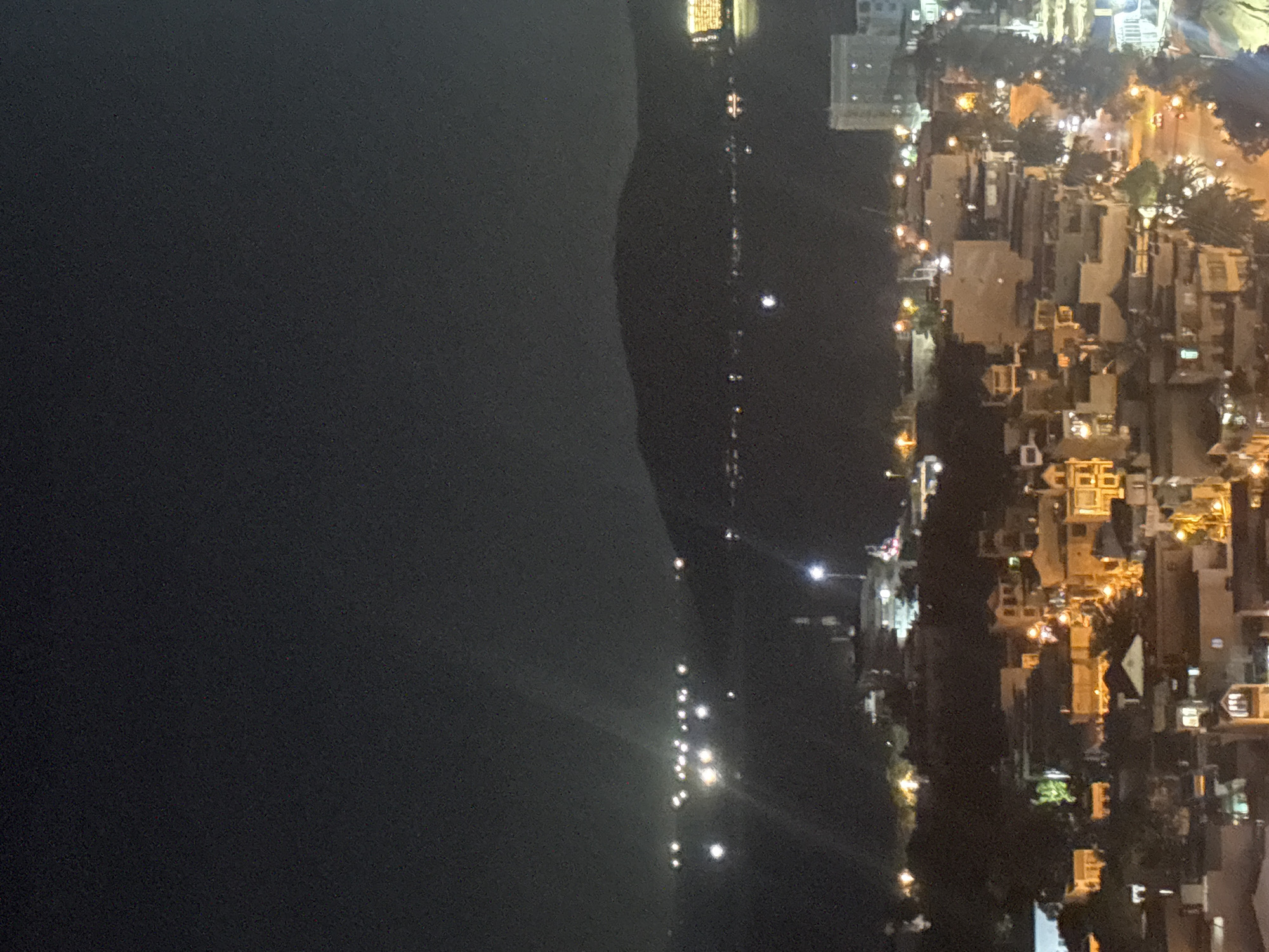 This is a night-time aerial photograph of the city of Nha Trang, Vietnam. The image captures a coastal city scene with a body of water in the background, likely Nha Trang Bay, and a mountainous silhouette beyond it. In the foreground, the city is illuminated by numerous lights from buildings, streetlights, and possibly vehicles, creating a warm glow against the dark sky. The architecture appears dense, with many multi-story buildings clustered together, suggesting an urban environment. The lights on the water might indicate boats or buoys. The overall impression is a bustling city at night, viewed from an elevated perspective.
