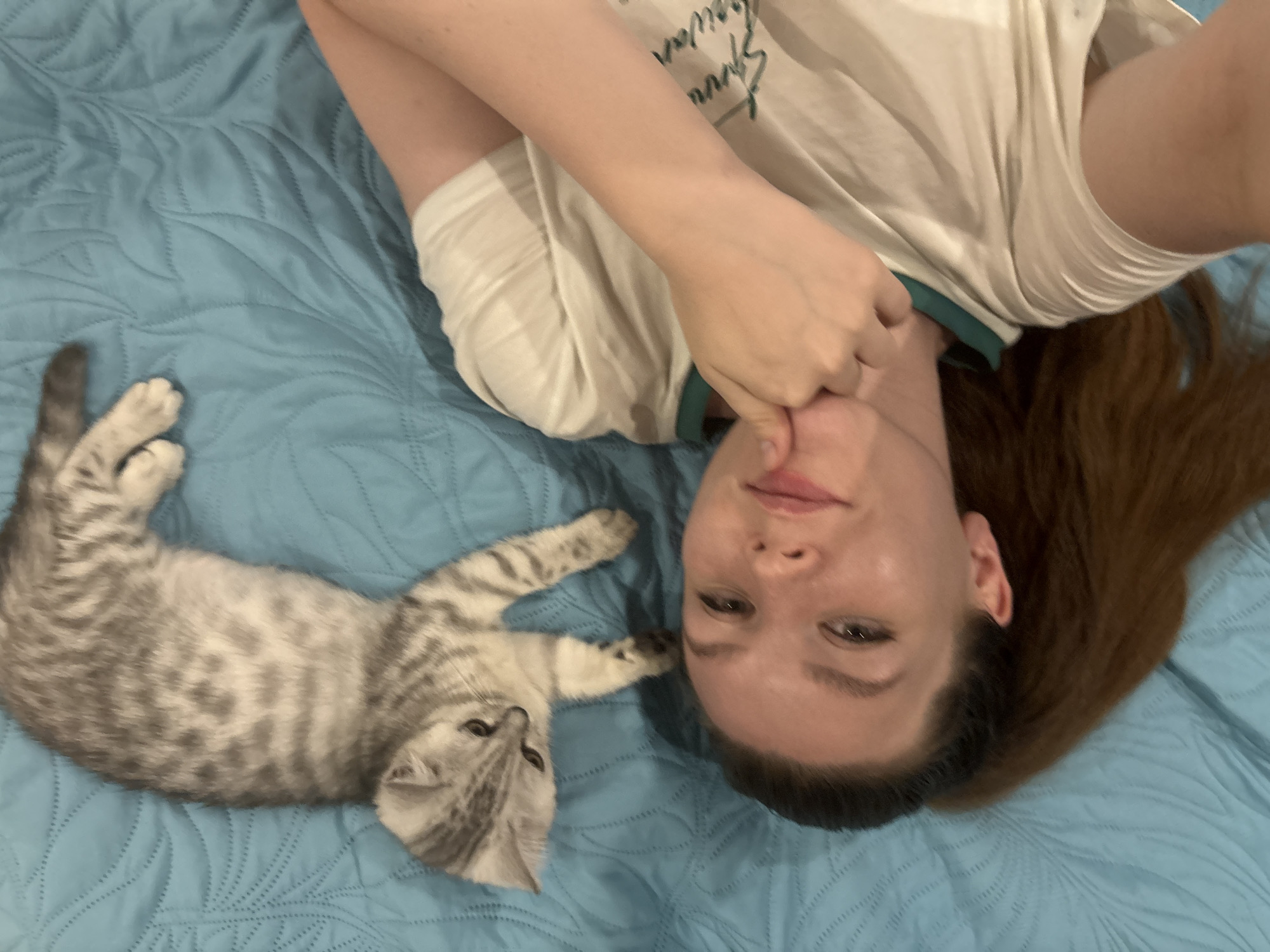 A light-skinned person is lying on a blue, quilted bedspread, oriented with their head towards the bottom of the frame. The person's brown hair is spread on the bed. The person is wearing a white shirt with dark green trim around the collar. Their left hand is raised near their face. A grey tabby kitten is lying on the bedspread to the left of the person. The kitten's head is oriented towards the bottom left corner of the frame. There is no information to determine the location in the city of Izhevsk, Russia.