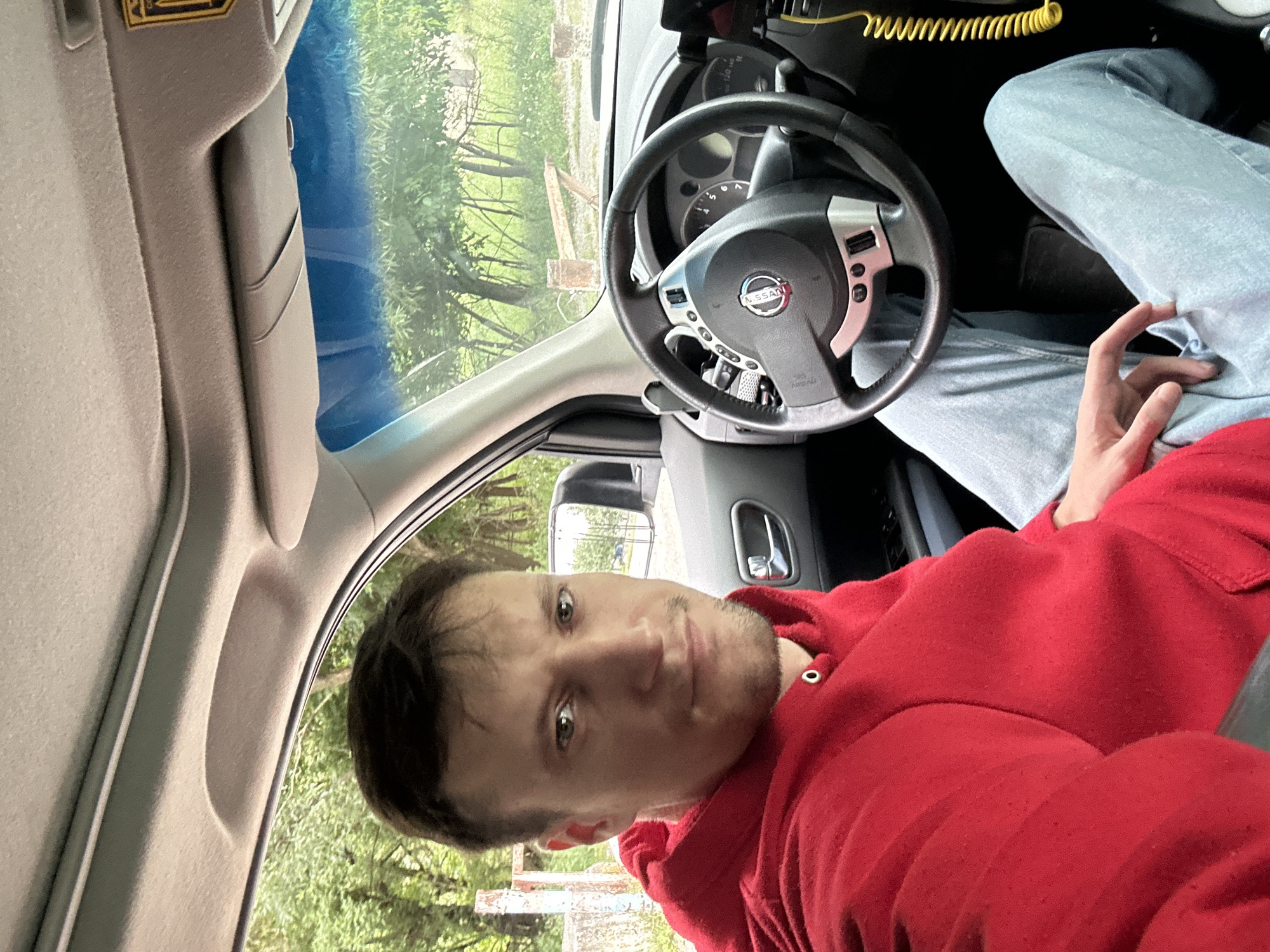 A medium shot captures a man sitting in the driver's seat of a car. He is looking directly at the camera with a neutral expression. The man is wearing a bright red hooded sweatshirt and light blue jeans. The interior of the car is visible, including the steering wheel, dashboard, and part of the passenger side door. A yellow coiled cord hangs from the dashboard. Outside the car, trees and greenery are visible through the windows, suggesting an outdoor setting, possibly on the outskirts of Makiv, Ukraine, given the presence of rustic wooden posts and fences in the background. The lighting suggests it is daytime.