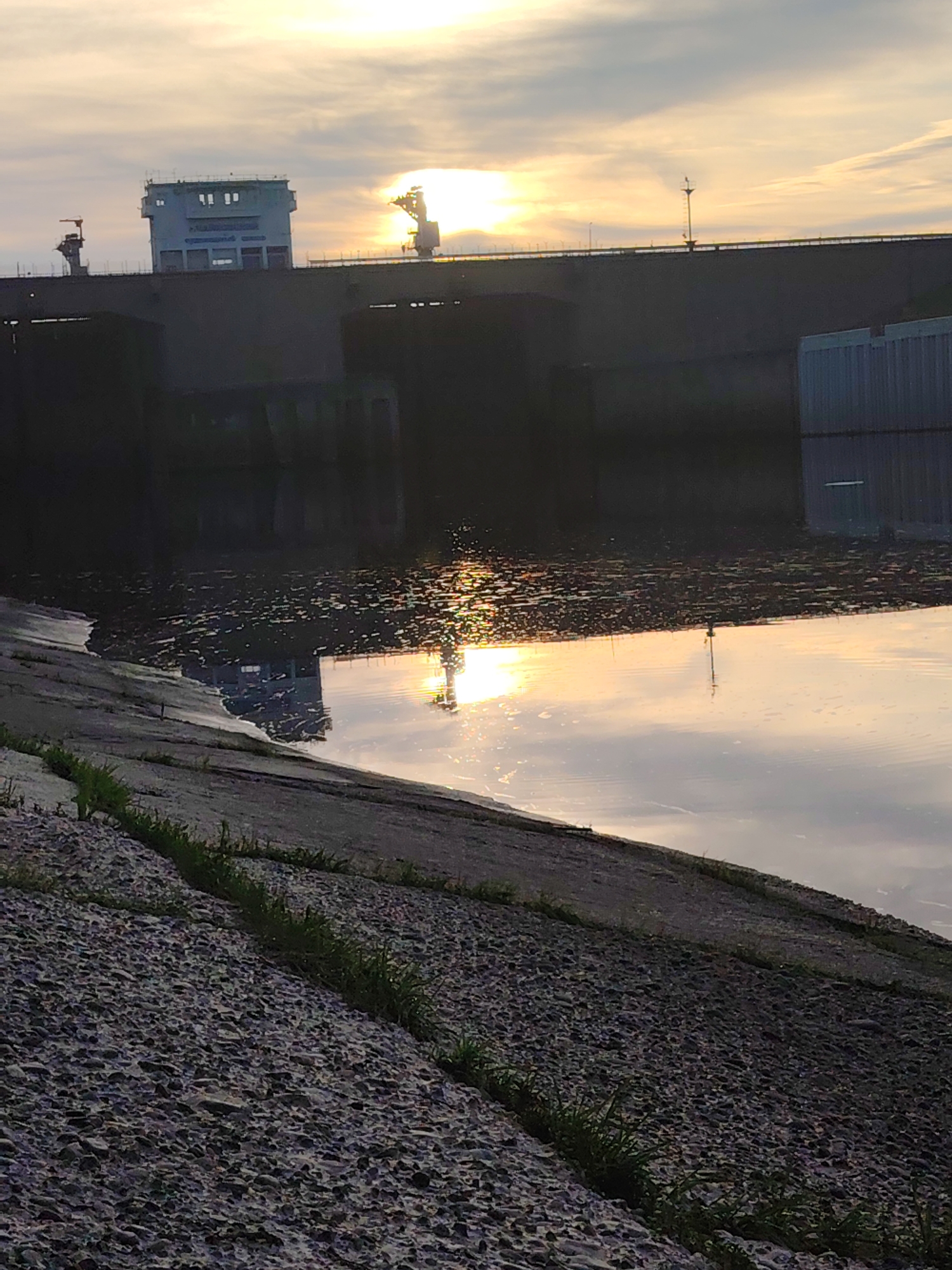 The image depicts a tranquil sunset or sunrise scene over a large body of water, likely a river, with prominent industrial infrastructure in the background. The setting suggests a dam or hydroelectric power station, consistent with the Chaikovsky, Russia context.
In the background, a massive, dark dam-like structure spans the width of the frame. Perched on top of this structure, a light-colored, multi-story building, possibly a control tower or administrative facility, stands out with several window-like openings. To its right, a tall industrial crane or similar machinery is silhouetted against the bright sky. A bridge or roadway, with faint indications of lampposts, extends across the top of the dam.
The sky, indicating either dawn or dusk, displays warm hues of orange and yellow near the horizon, transitioning to lighter blues and greys overhead. The sun is partially obscured by the background structures, casting a strong glare that creates shimmering reflections on the calm water surface below.
The water in the mid-ground reflects the bright sky and the central building, creating a distorted yet recognizable mirror image. Patches of floating debris or ripples are visible on the water's surface, particularly where the sunlight hits.
In the foreground, a sloped bank constructed from rough, dark grey material, possibly concrete slabs or reinforced stone, extends from the lower left towards the mid-right. Small tufts of green grass grow sporadically in the cracks of this material.
No people or specific activities are visible, presenting a serene yet powerful landscape that combines natural beauty with human engineering at a transitional time of day. There is no visible text in the image.