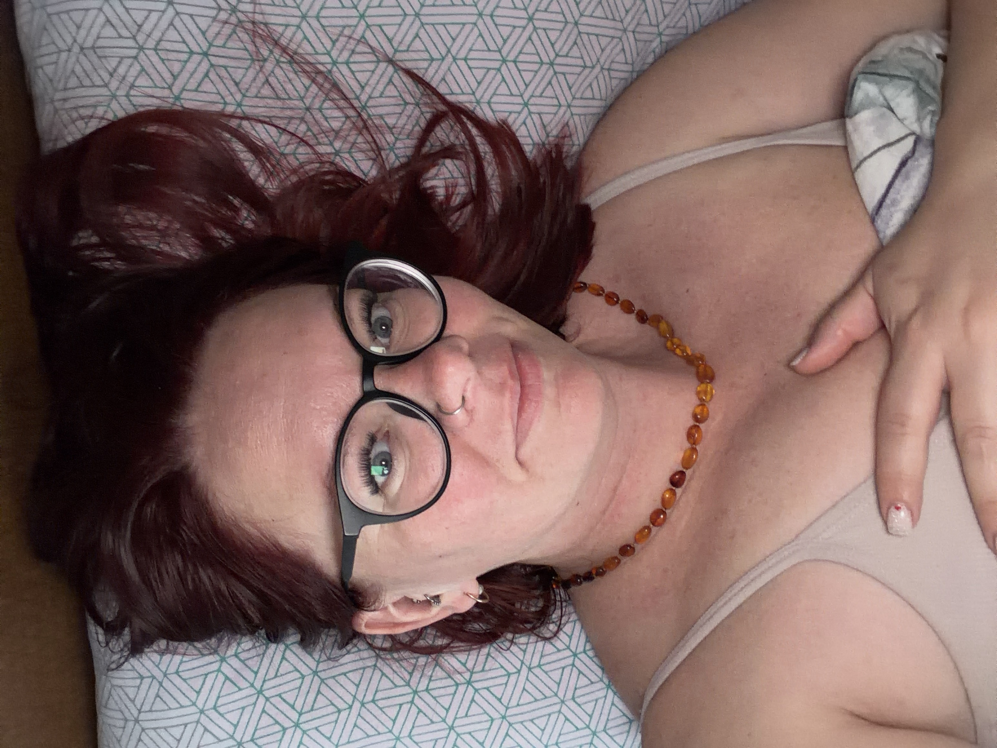 A woman with red hair and glasses lies on a bed. She wears a tan-colored top and an amber necklace. Her right hand is near her chest. The bed has a white and green patterned sheet. The image does not provide information to determine that it was taken in the city of Chaikovsky, Russia.