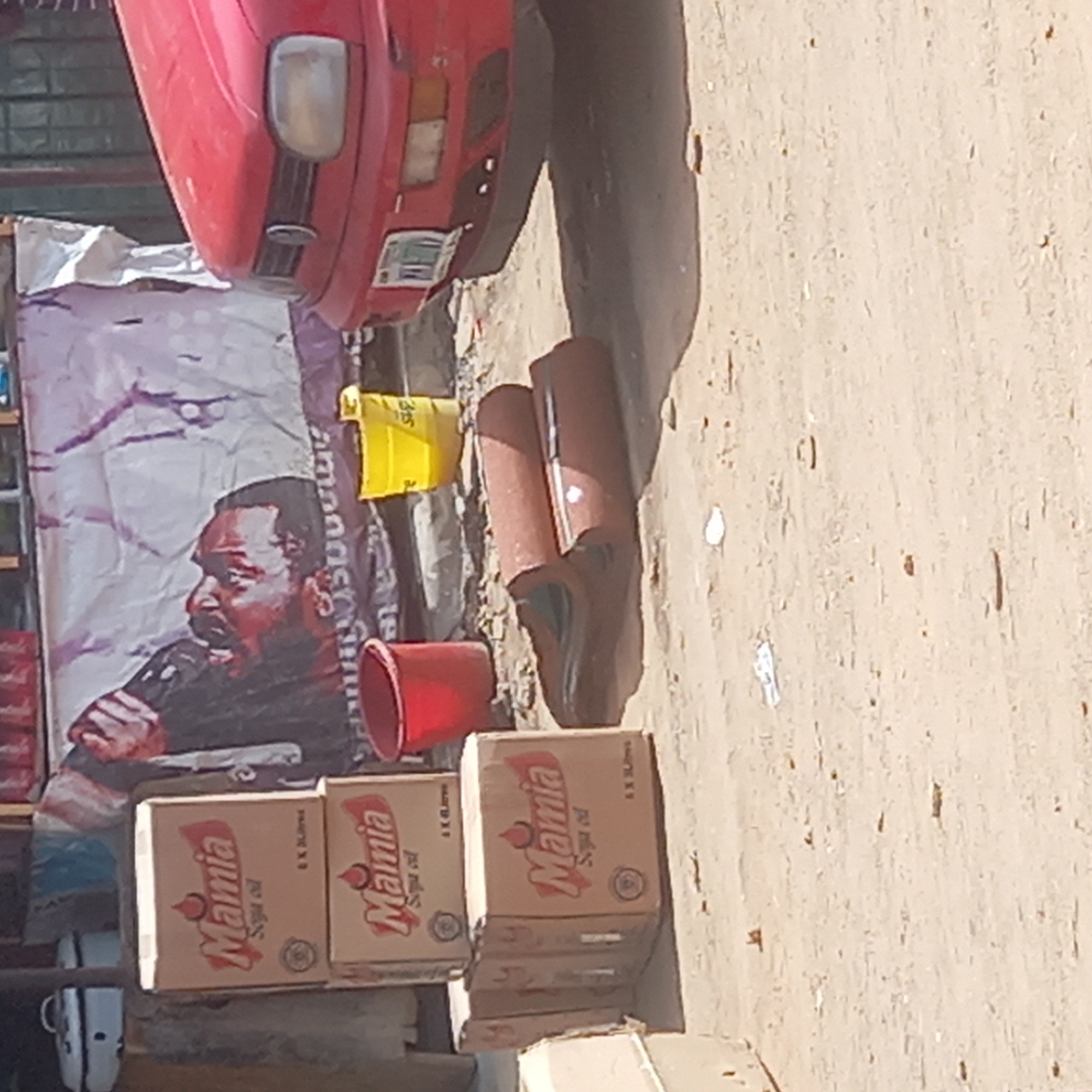This image captures an outdoor street or market scene, likely in Dadi Riba, Nigeria, characterized by bright daylight and a dusty, light-colored ground.
In the foreground and midground, several cardboard boxes are stacked. These boxes prominently feature the brand name "Mamia Soya oil" and indicate a quantity such as "8 X 1L" or "8 X 1.5L" (visible as "AX BLI" in OCR). A red plastic bucket and a yellow plastic bucket, marked with "365", are also placed on the ground. Nearby, several curved, brown objects, possibly ceramic roof tiles or drainage pipes, are scattered, with a pair of brown shoes resting among them. A white mug with black spots is partially visible on the far left, near the bottom.
To the left, a large banner or poster is displayed, featuring a headshot of a man with a dark complexion and a beard, appearing to speak into a microphone. Parts of text like "smmmis" and "ate" are visible on this banner. The upper portion of the image shows the front-right section of a red car, including its headlight and part of the bumper.
The scene is static, depicting various goods and objects arranged outdoors, suggesting a commercial setting such as a roadside stall or market. The items appear somewhat dusty, consistent with an open-air environment.
