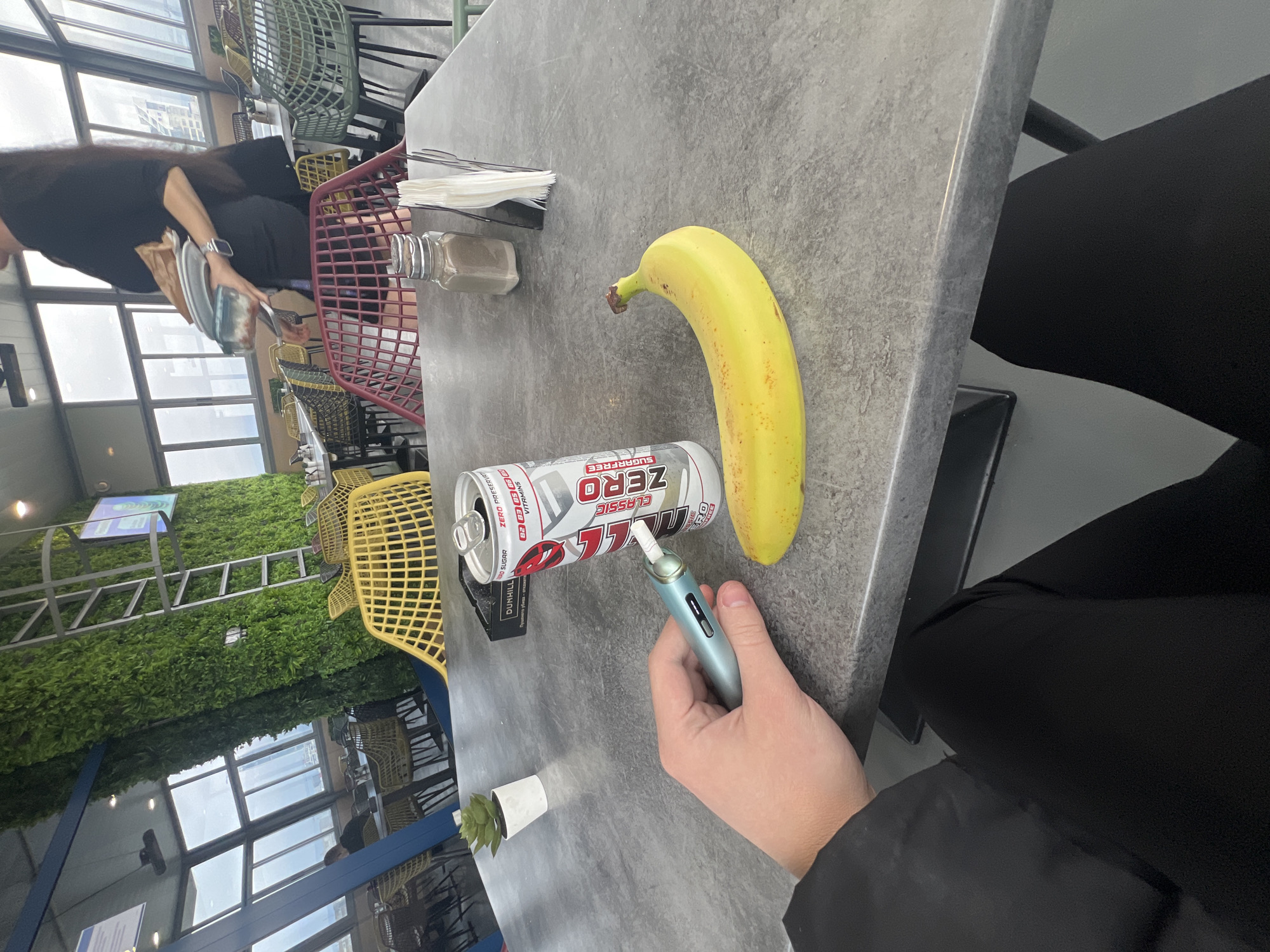 The image shows a gray table with a banana, a can of an energy drink, a blue electronic device, and a hand holding the device. There's a small plant in a white pot near the edge of the table. In the background, there are chairs, a green wall, and a view of a cityscape through large windows. A person with dark hair is sitting at the table, and a red shopping basket is on the table.
