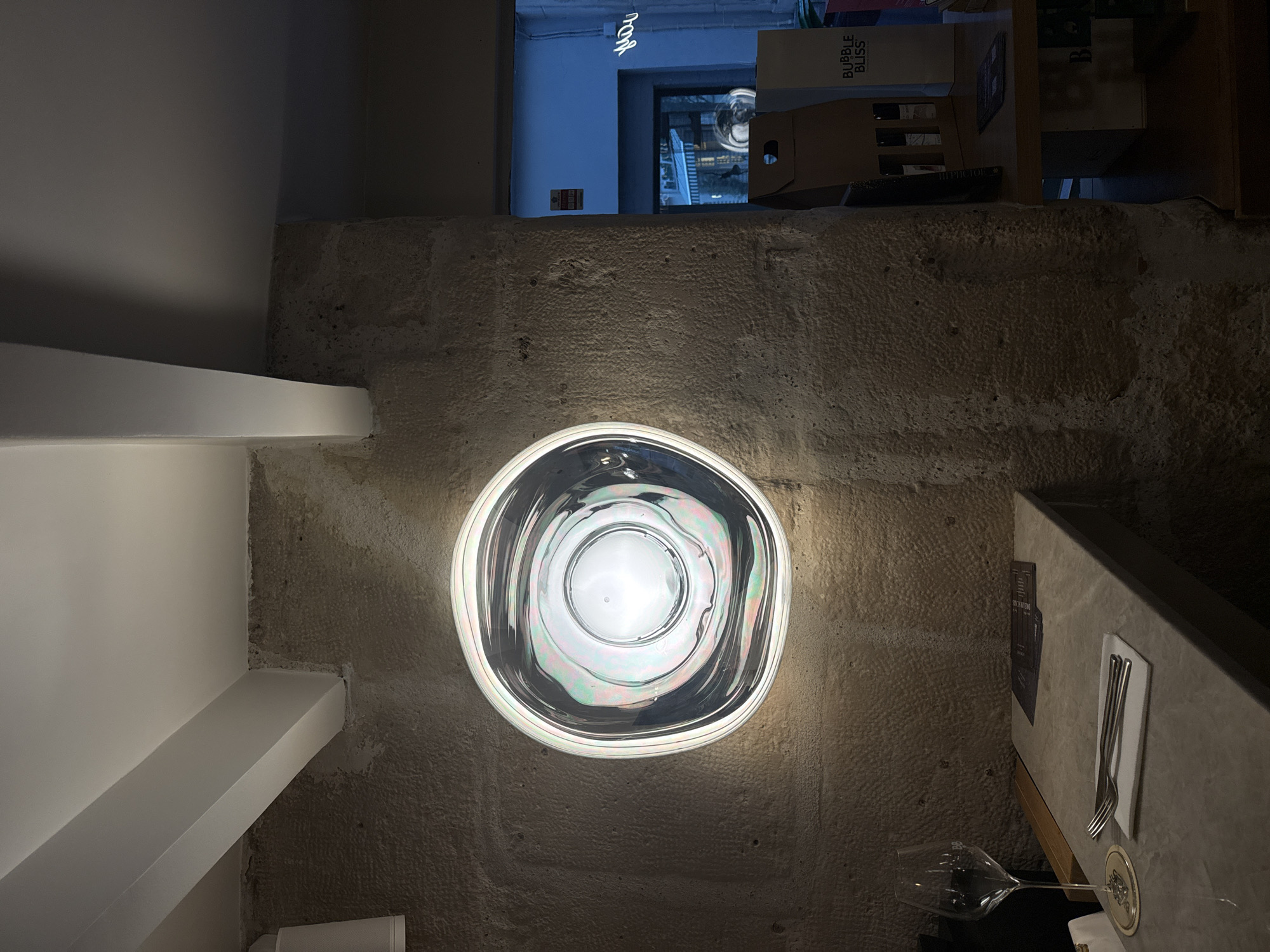 The image shows an interior shot of a restaurant or dining space in Paris, France. The location is suggested by the architectural style and possibly the "BUBBLE BLISS" packaging visible in the background near the upper edge.
The foreground features a rough, textured stone wall with an unusual, round, illuminated wall fixture. The light fixture has an iridescent, almost liquid-like appearance. To the left of the wall, there's a section of a white, modern-looking beam or architectural element.
To the right, a dining table is visible. It has a menu card and a place setting that includes cutlery. A wine glass is placed near the edge of the table.
In the background, above the stone wall, there is a glimpse into what appears to be another area or space, possibly an entryway. There is a box labelled "BUBBLE BLISS" and other objects suggesting storage or retail display space. A sign is visible in the upper left with a handwritten, illuminated script. The background lighting is a cooler blue tone.