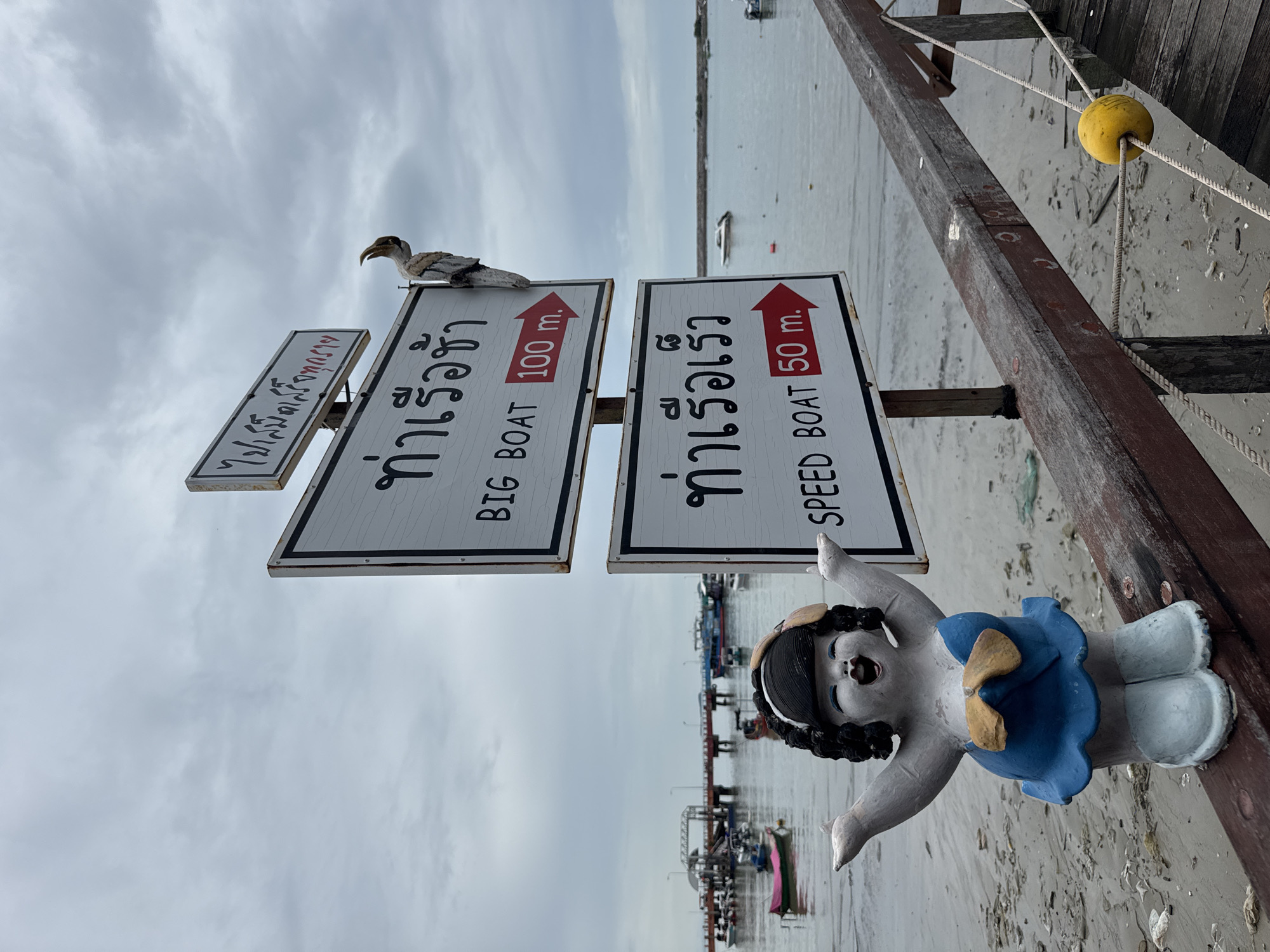 The image shows a pier in Ban Phe, Thailand, on an overcast day. In the foreground, a ceramic figurine of a woman with black curly hair, wearing a blue dress and white boots, stands on a wooden railing. To the right of the figurine are two white directional signs with black lettering. The top sign, angled slightly, has Thai script and reads "BIG BOAT 100 m." with a red arrow. The bottom sign reads "SPEED BOAT 50 m." with a red arrow. A wooden bird sculpture is perched on the pole holding the signs. The pier extends into a body of water with several boats docked and anchored, including fishing boats and a larger passenger boat. The water is calm, and the shoreline is visible with sand and debris. A yellow buoy is attached to ropes on the wooden railing in the lower right quadrant of the image.
