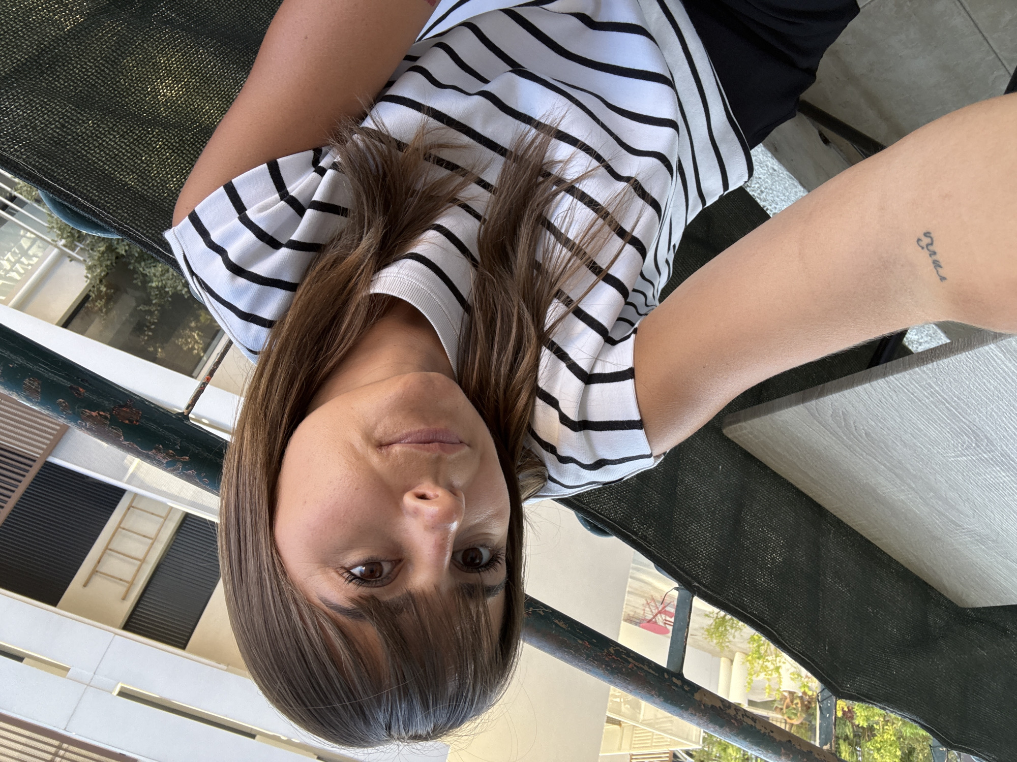 A person is seen from a low angle, likely a selfie, with their head at the bottom of the frame. The person has brown hair with bangs and is wearing a black and white striped shirt. The background includes a building facade, part of a green mesh structure with a metal frame, and a light-colored surface. The person's arm is visible, and a small tattoo is on the inner side of the arm. It's impossible to pinpoint the exact location to the city of Cannes, France, from the scene's features. The description reflects the objective visual elements without assumptions.