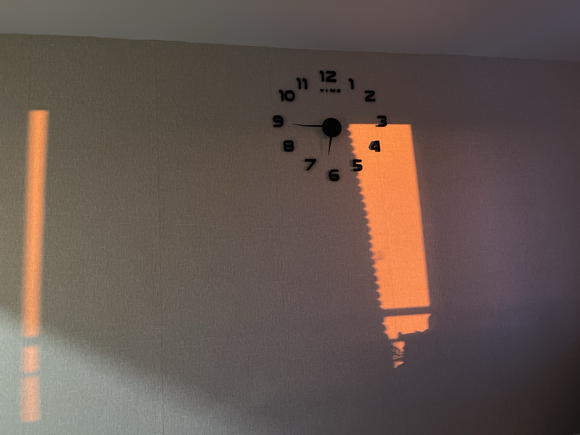 A wall clock is mounted on a textured wall. The clock has black numbers and hands. The hands indicate it is 9:31. Sunlight casts an orange glow near the clock. The lower part of the sunlight rectangle has a shadow cast from what appears to be window blinds.
This image may have been taken in the city of Chaikovsky, Russia. However, there is no way to definitively confirm this based on the content of the image.