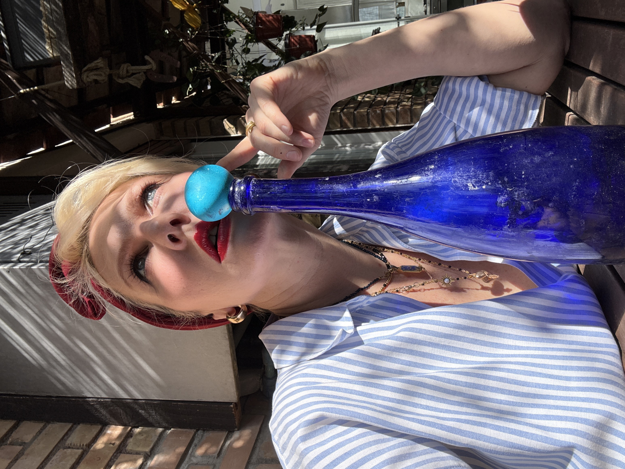 A woman with blonde hair and red lipstick poses with a blue glass bottle, the neck of which is pressed against her nose. She is wearing a blue and white striped shirt, gold earrings, a red headband, and layered necklaces. Her right index finger is pointing near the bottle's neck. The background shows a partially shaded outdoor setting, possibly a terrace or garden, with plants and building structures. The exact location in Rome, Italy, cannot be determined from the image alone.