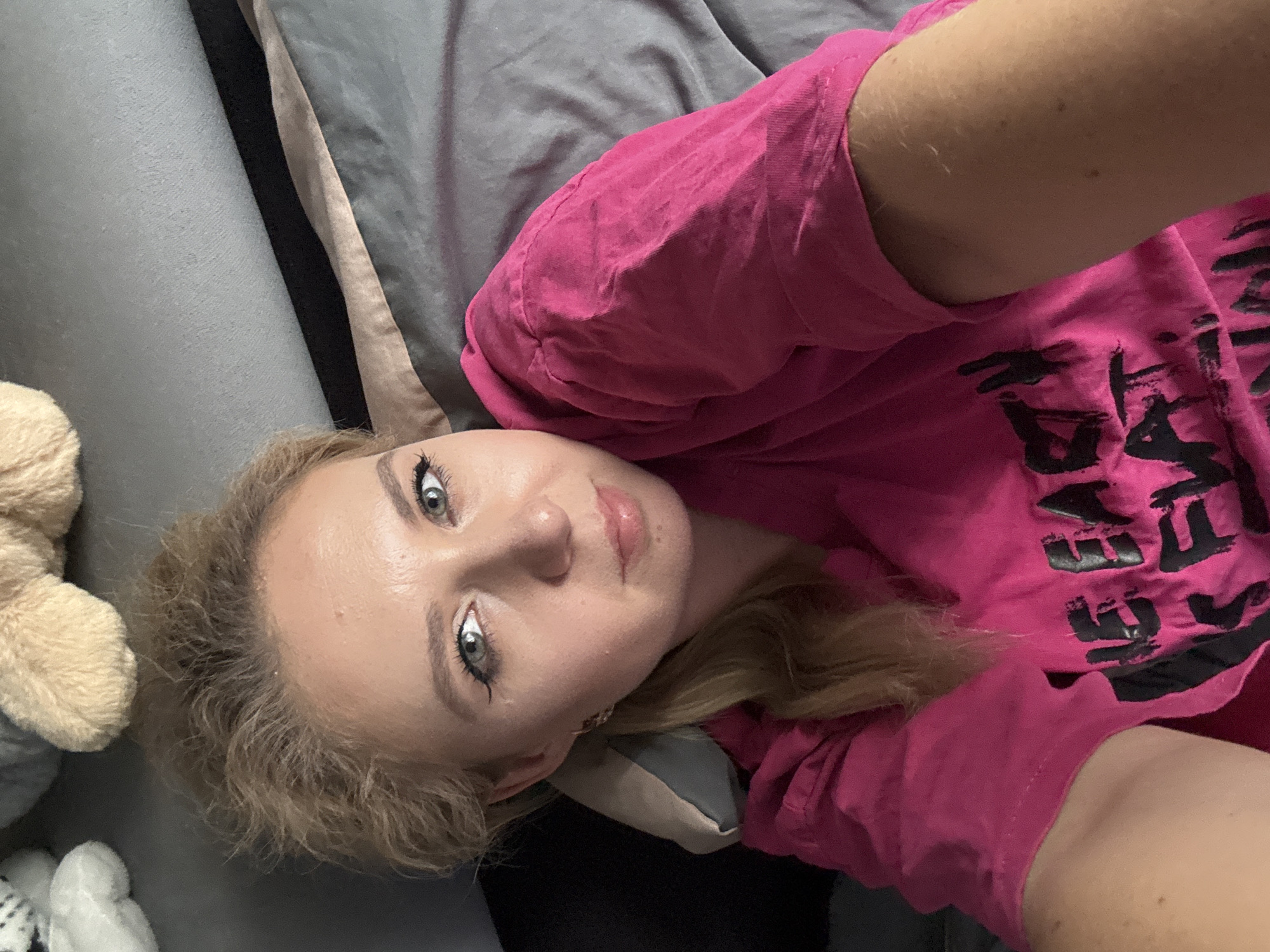 A woman with blonde hair is lying on a grey couch, looking at the camera. She is wearing a bright pink t-shirt with black lettering that reads "THE EARTH IS FLAT." There are stuffed animals in the background. The image is an indoor selfie taken from a high angle.