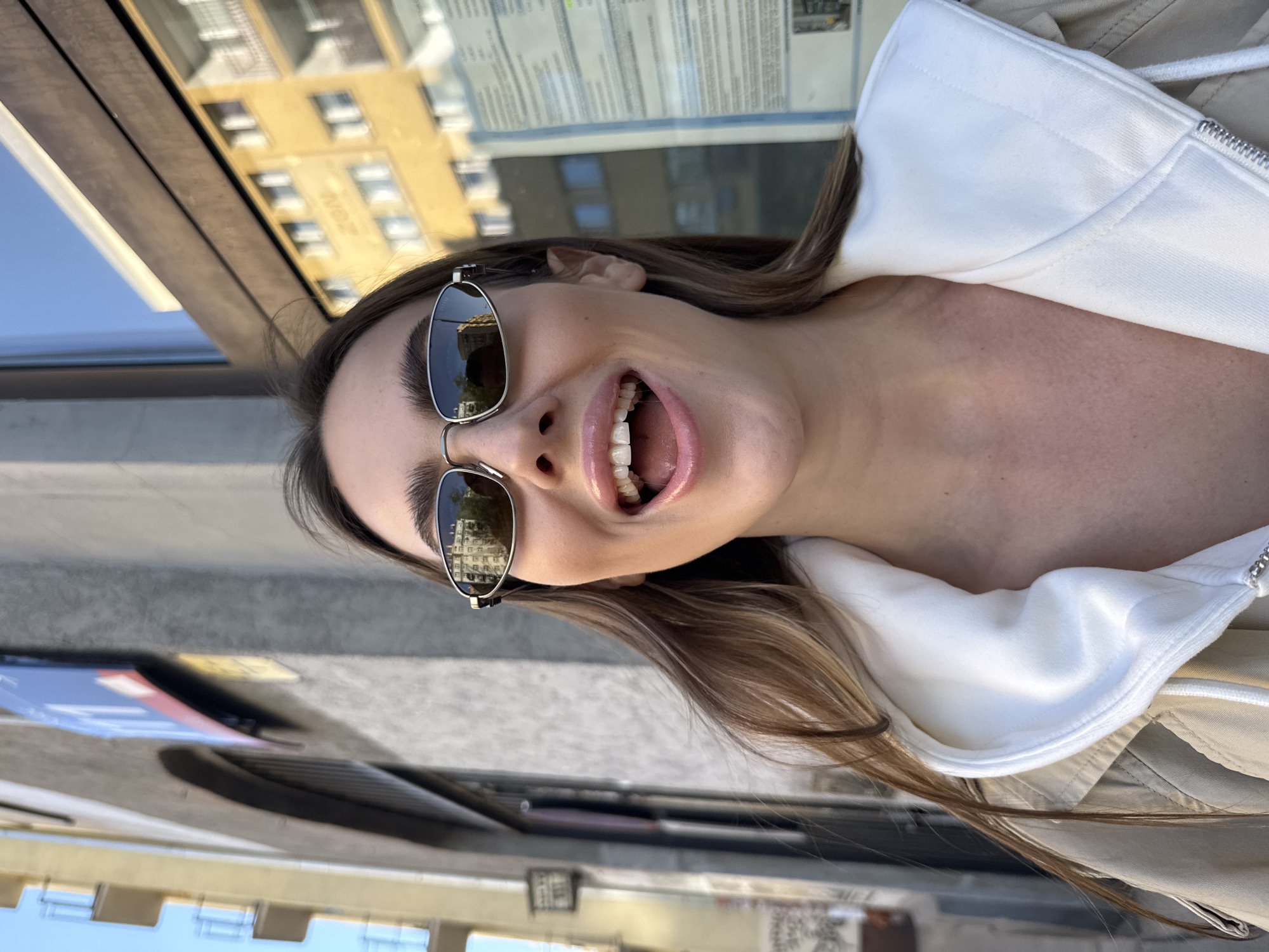 A young woman wearing sunglasses and a white hoodie stands outside in Warsaw, Poland, appearing to smile or laugh with her mouth open. The reflection in her sunglasses suggests she is near buildings, consistent with the urban setting. The environment shows elements of street furniture or signage, indicating a public area, potentially a sidewalk café or restaurant.