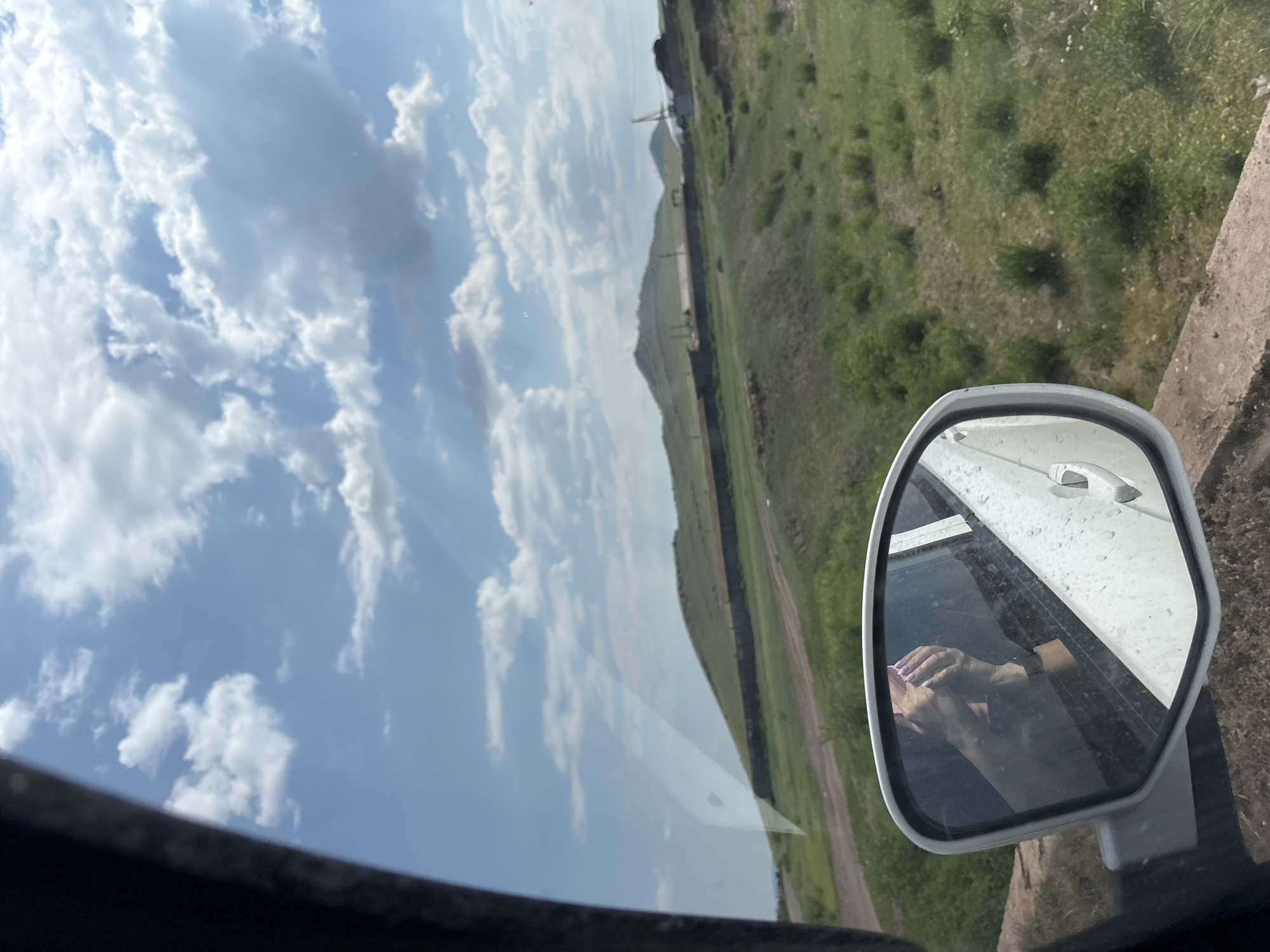 The image captures a view from inside a vehicle, looking out through a side window and a side-view mirror. The mirror reflects two hands, one on top of the other, with purple nail polish visible on the fingernails. The background visible through the car window is a rural landscape under a partly cloudy sky. Gentle, rolling hills covered in green grass and sparse vegetation dominate the mid-ground, with a dark, linear structure running along the base of the nearest hill. In the distance, there are some buildings and what appears to be construction equipment or industrial structures. The foreground shows the side of the car, including part of the door and the exterior mirror housing, which appears to be made of a textured material. The sky is a bright blue with scattered white clouds. The overall impression is of a drive through a rural area. The provided location of Troitskoye, Russia, suggests a setting in the Russian countryside.