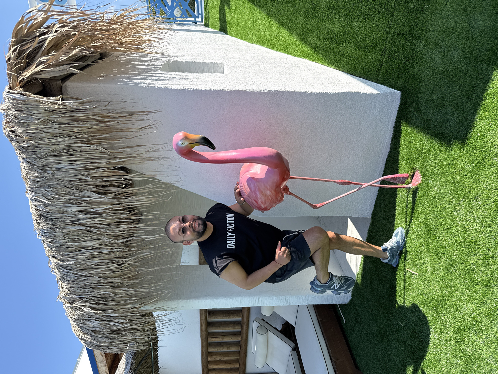 In Baliovtsi, Bulgaria, a man poses with a pink flamingo statue in front of a white building with a thatched roof. The man is wearing a black t-shirt that says "DAILY FICTION," black shorts, and grey sneakers. He is smiling and has one leg bent at the knee. The flamingo statue is positioned to his right, and it is approximately the same height as the man. The building appears to be a small structure, possibly a beach hut or cabana, with a rough white exterior and a thatched roof made of dried palm leaves. Artificial grass covers the ground, creating a vibrant green surface. In the background, lounge chairs are visible, suggesting a relaxation area. The sky is clear and blue, indicating a sunny day.
