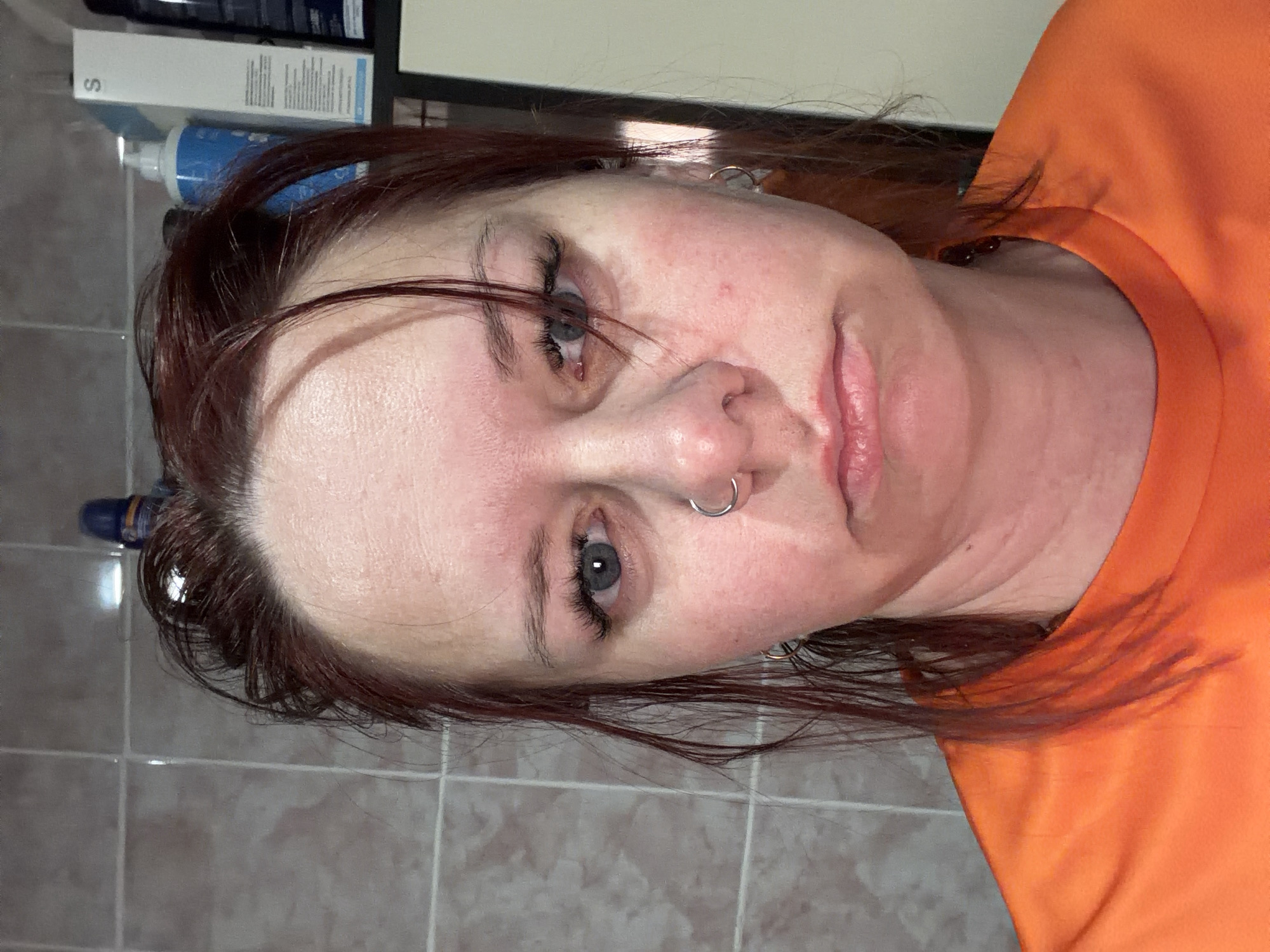 A close-up portrait photograph features a fair-skinned woman with light blue eyes and reddish-brown hair. She has a nose ring and earrings. She is wearing an orange T-shirt. The background suggests a bathroom setting with tiled walls and cosmetic bottles in the upper left corner. The image does not contain information relating to the city of Chaikovsky, Russia.