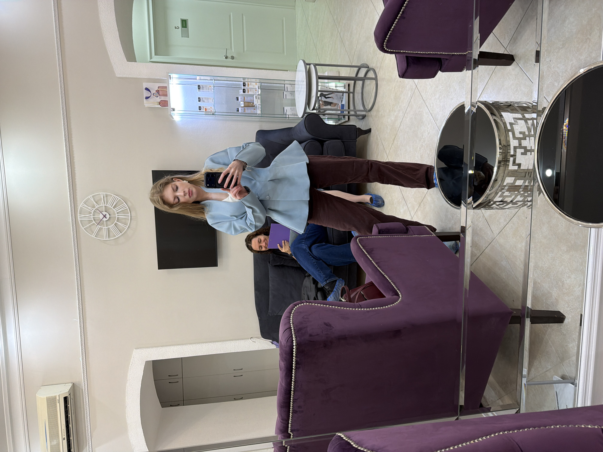 The image depicts an interior scene, likely a waiting area or lobby. A blonde woman is posing for a selfie in front of a wall mirror. She is wearing a light blue jacket and brown pants. Behind her, a woman sits on a dark-colored sofa, holding a purple folder. The waiting area contains purple armchairs, a black television on the wall, a clock, and a display case with various items. The floor is tiled. The location is possibly in Moscow, Russia.