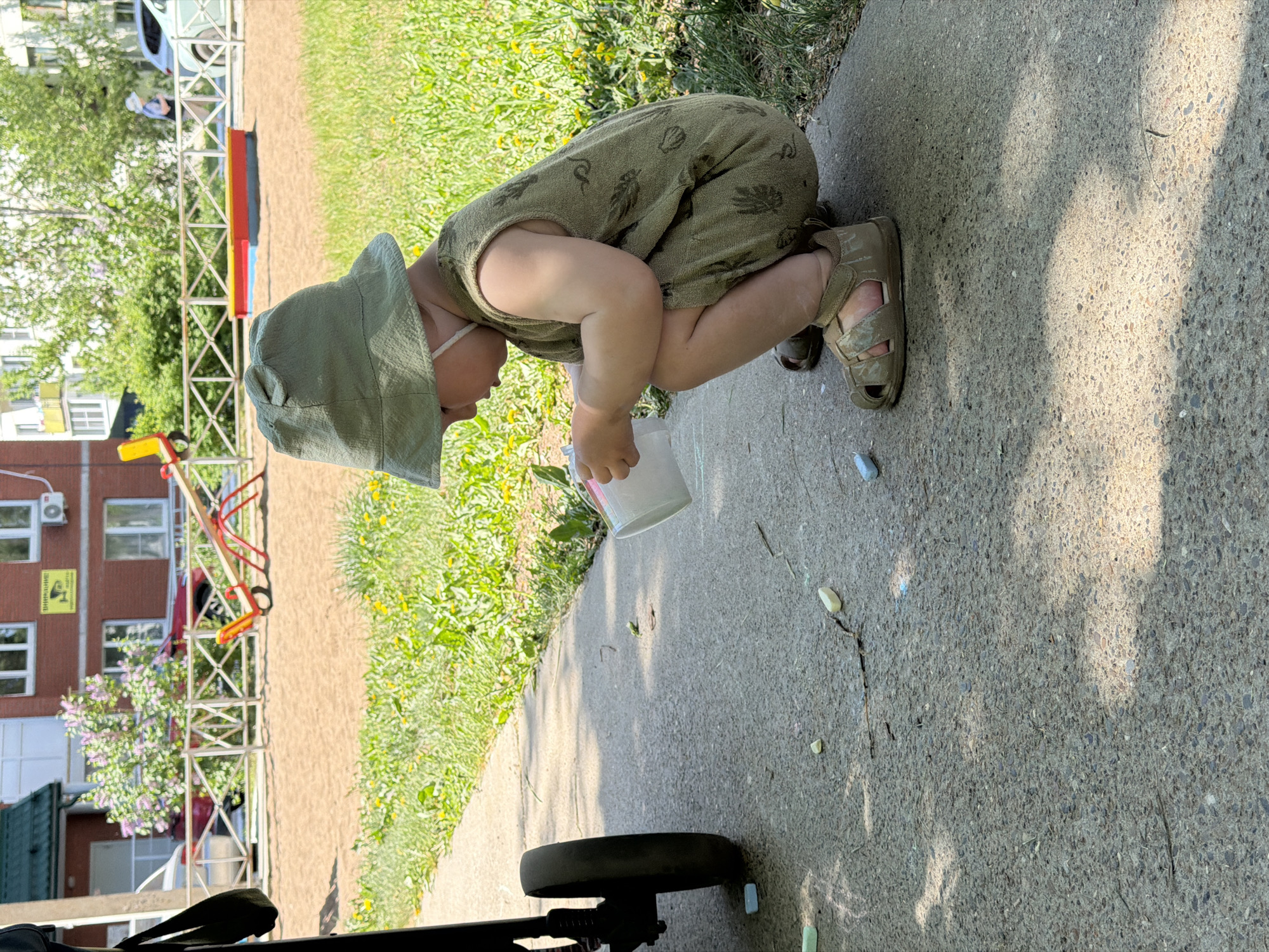 A young child wearing a green sunsuit and matching hat is crouched down on a concrete sidewalk, holding a white cup. The child appears to be interacting with the sidewalk, possibly drawing with chalk, as several pieces of chalk are visible nearby. The sidewalk is adjacent to a grassy area with dandelions. In the background, a playground and a building can be seen, potentially identifying the location as a park or residential area in Chaikovsky, Russia. A stroller wheel is visible in the foreground.