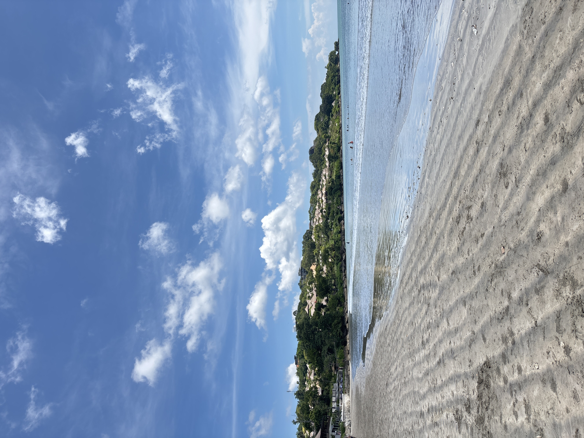 The image depicts a beach scene in Kuta Selatan, Indonesia. The camera points out to the ocean. The beach is on the right side of the photo and is sandy and ridged with water from the ocean covering part of the sand. The ridge of land beyond the ocean appears covered in lush green trees and vegetation. Buildings appear nestled amongst the trees. The sky is blue with scattered puffy white clouds. Some people appear to be in the water, in the distance.