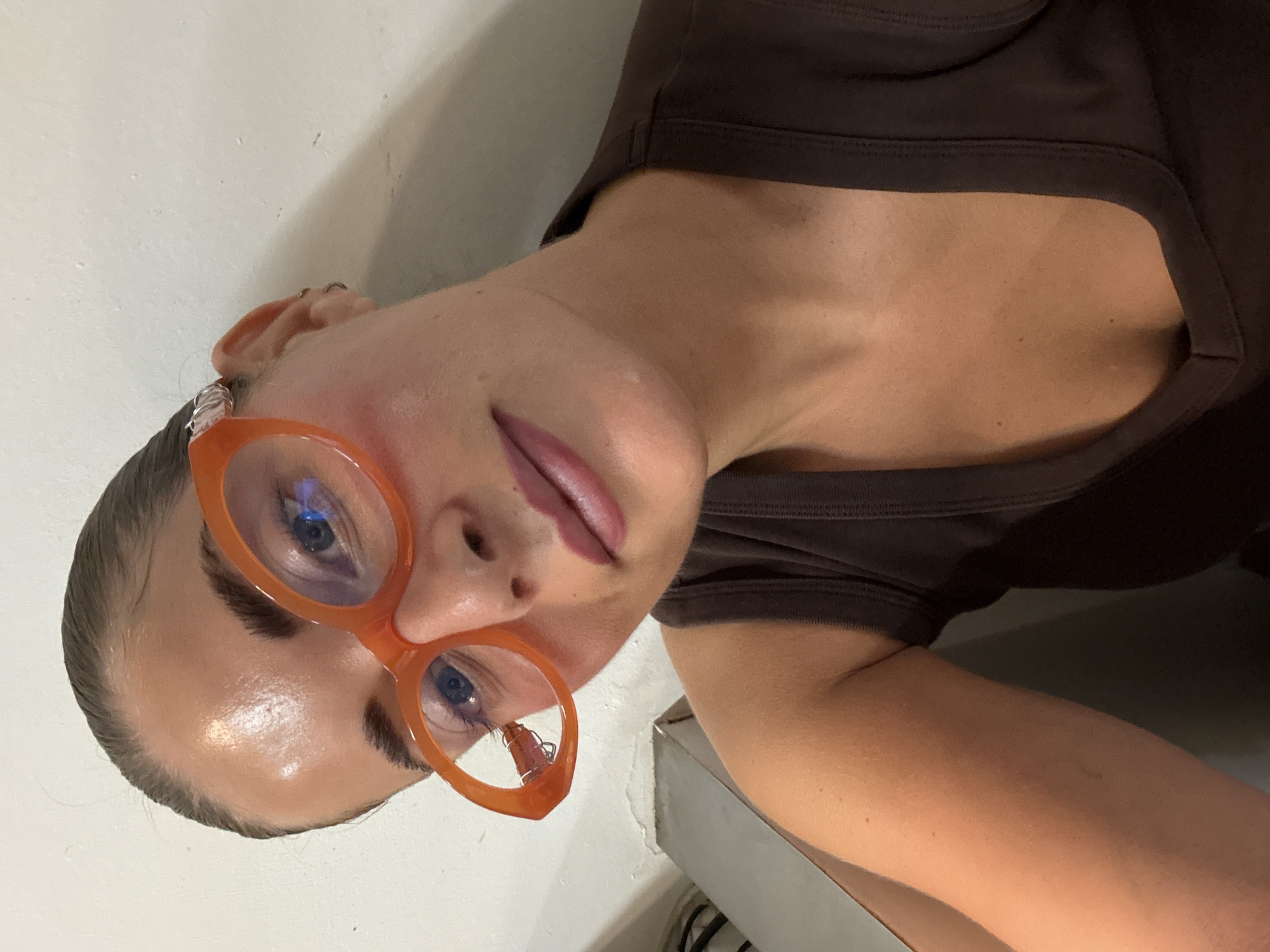 This is a close-up, eye-level, indoor selfie of a woman with tan skin and dark hair pulled back. She is wearing large, orange, oval-shaped glasses. The glasses have a silver decorative element on the left temple. She is also wearing a dark brown tank top. Her makeup includes blush on her cheeks and lipstick. The background is a plain, light-colored wall with a shadow on the right side. A small portion of a grey metal object is visible in the lower left corner. The lighting appears to be artificial. There is no discernible information about the location in Paris, France.