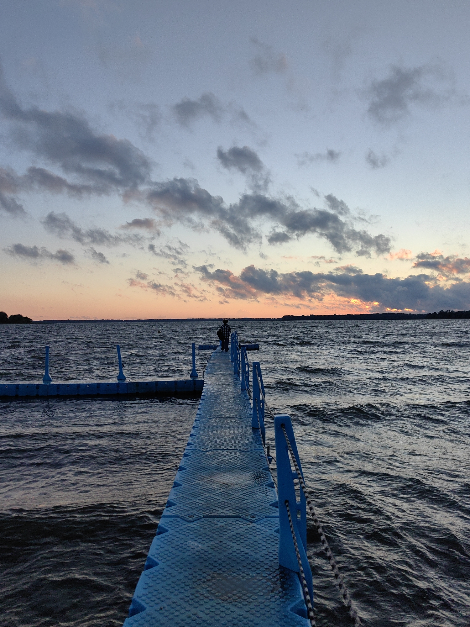 A person wearing a checkered jacket stands at the end of a blue floating dock extending into a dark, choppy body of water under a cloudy sunset sky. The sky displays hues of orange, pink, and blue with scattered gray clouds. The water is rippled by wind. The dock has blue railings and a textured surface. In the distance, a treeline is visible along the horizon. The image appears to be taken in Myadzyel, Belarus, likely at Lake Myastro or a similar large body of water near the town.