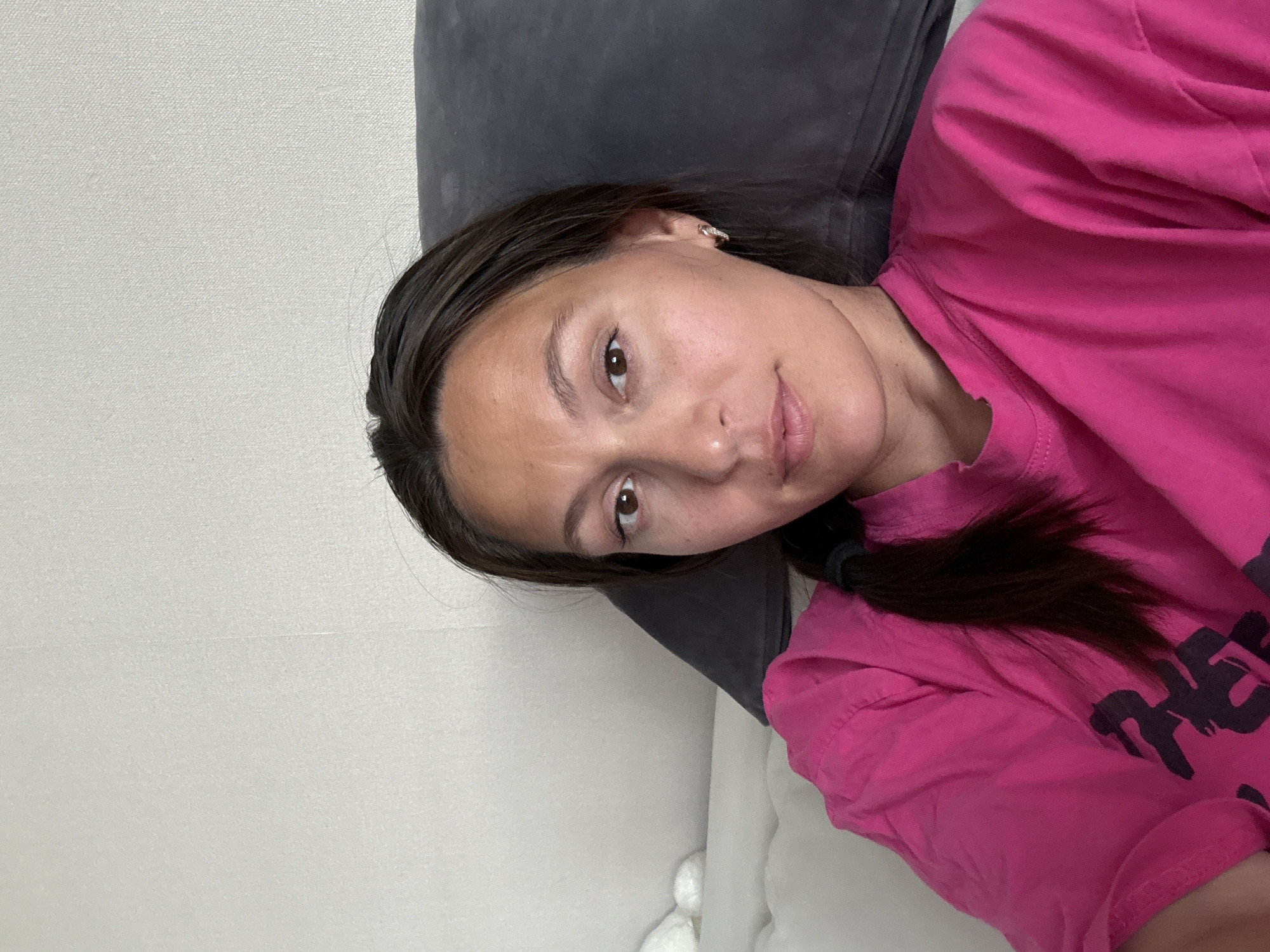 The image shows a woman lying down, resting her head on a dark grey pillow. She is wearing a pink t-shirt. Her dark hair is pulled back in a ponytail. The background consists of a light grey wall. The location is unknown, it is not clear if the location is Chaikovsky, Russia.