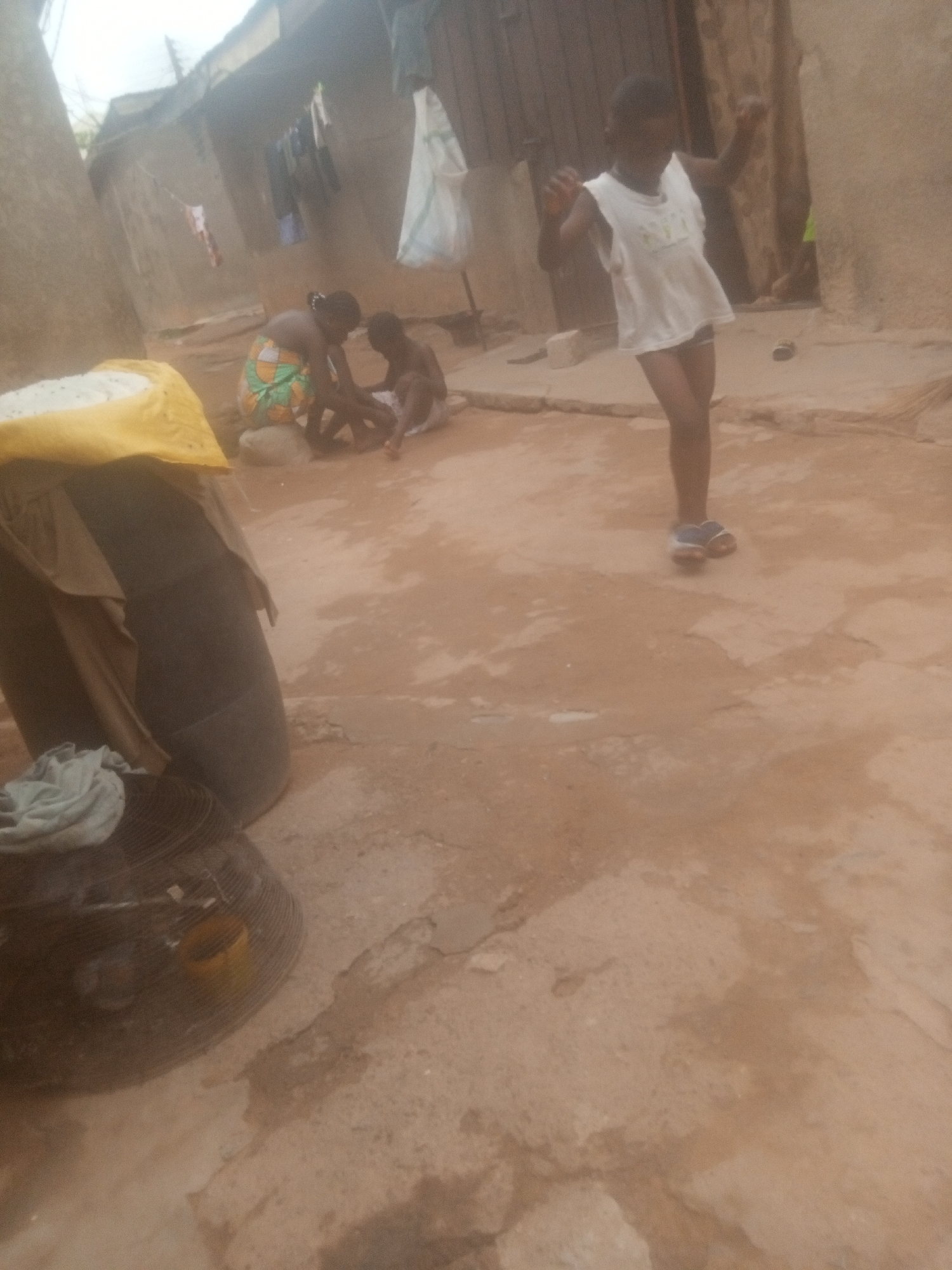 The image depicts an outdoor scene, possibly a courtyard or yard, with a reddish-brown dirt surface. In the background, two individuals are seated on the ground, one possibly attending to the other's foot. Further back, a child in a white tank top stands with their arms raised. To the left of the frame, objects including what appears to be a black tire, a wire structure, and items covered with fabric are present. Laundry hangs on a line in the background. The location is in a rural area of Unknown, Unknown.