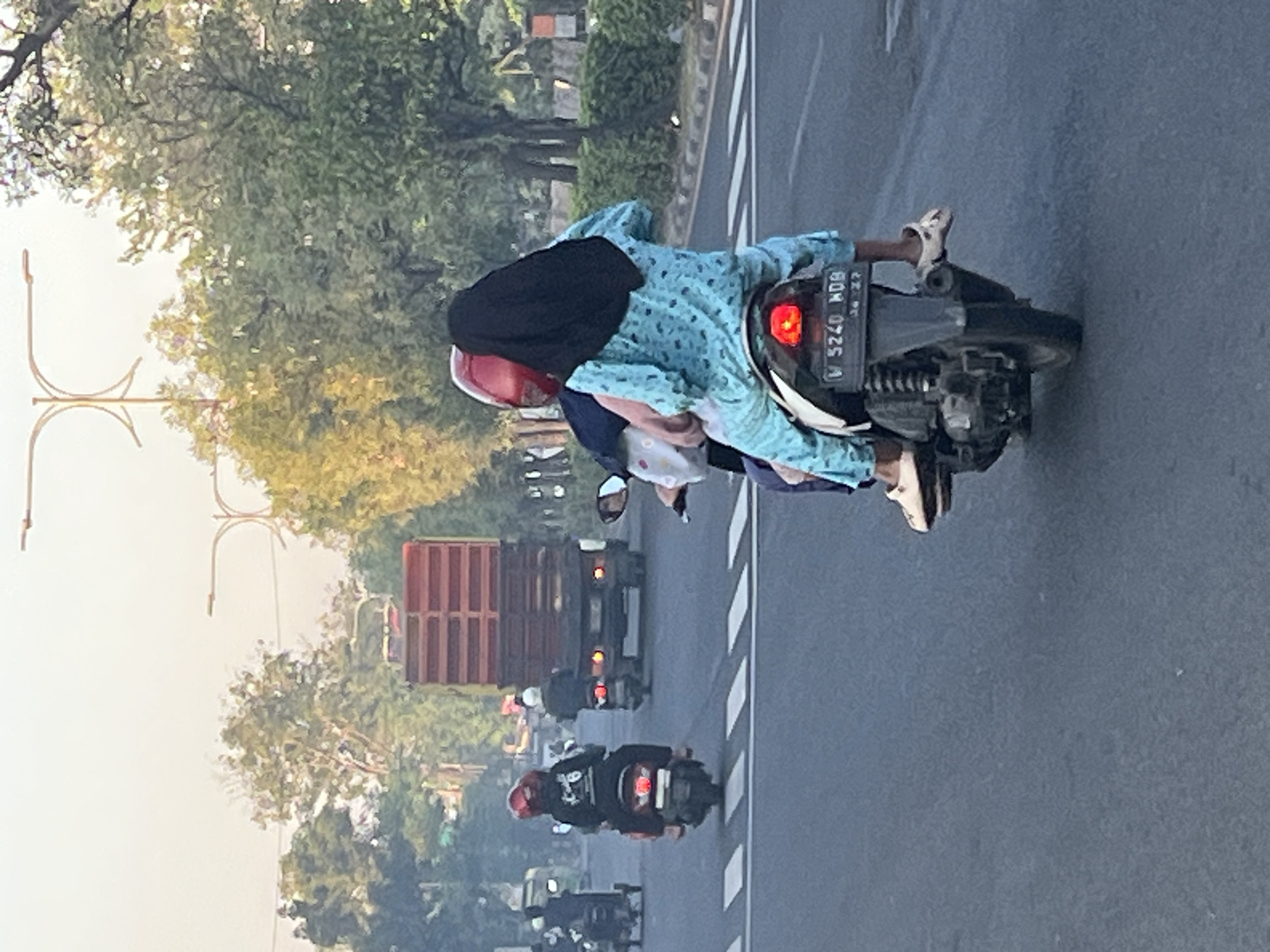 In Kludan, Indonesia, on a paved street, two individuals ride on a motorcycle. The person in the front wears a red helmet and a dark shirt. The person behind them wears a blue patterned shirt and a black head covering, with a baby or child held between them. The motorcycle is dark colored and has a visible license plate reading "W 5240 MDB". The road has white pedestrian crossing lines. In the background, a large red truck is visible, as are trees and utility poles. The sky is hazy and appears to be either early morning or late afternoon.