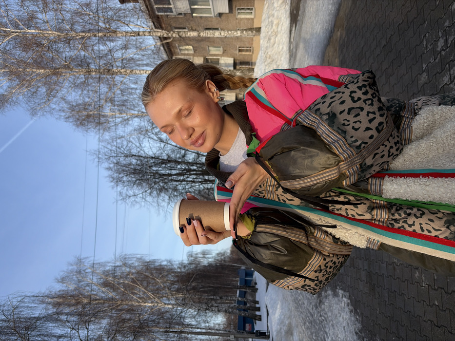 A fair-skinned individual, likely female, with blonde hair pulled back, stands outdoors in Chaikovsky, Russia, on a bright, sunny day. The person is oriented towards the right, with their eyes closed and face angled upward, appearing to enjoy the sunlight. They are wearing a brightly colored, layered coat featuring sections of hot pink, olive green, leopard print, and white faux-shearling, with colorful striped accents and black drawstrings on the sleeves. A light grey t-shirt is visible underneath. The person holds a plain brown paper coffee cup with a white lid in both hands, presenting it slightly forward. Their fingernails are painted dark and light pink.
The environment suggests late winter or early spring. A paved pedestrian walkway, partially covered with patches of melting snow, forms the foreground. In the background, tall, bare deciduous trees, possibly birch, line what appears to be a street or park area. A multi-story brick residential building with multiple windows is visible on the right. Overhead, a clear blue sky is punctuated by a faint contrail from an aircraft and visible utility lines.