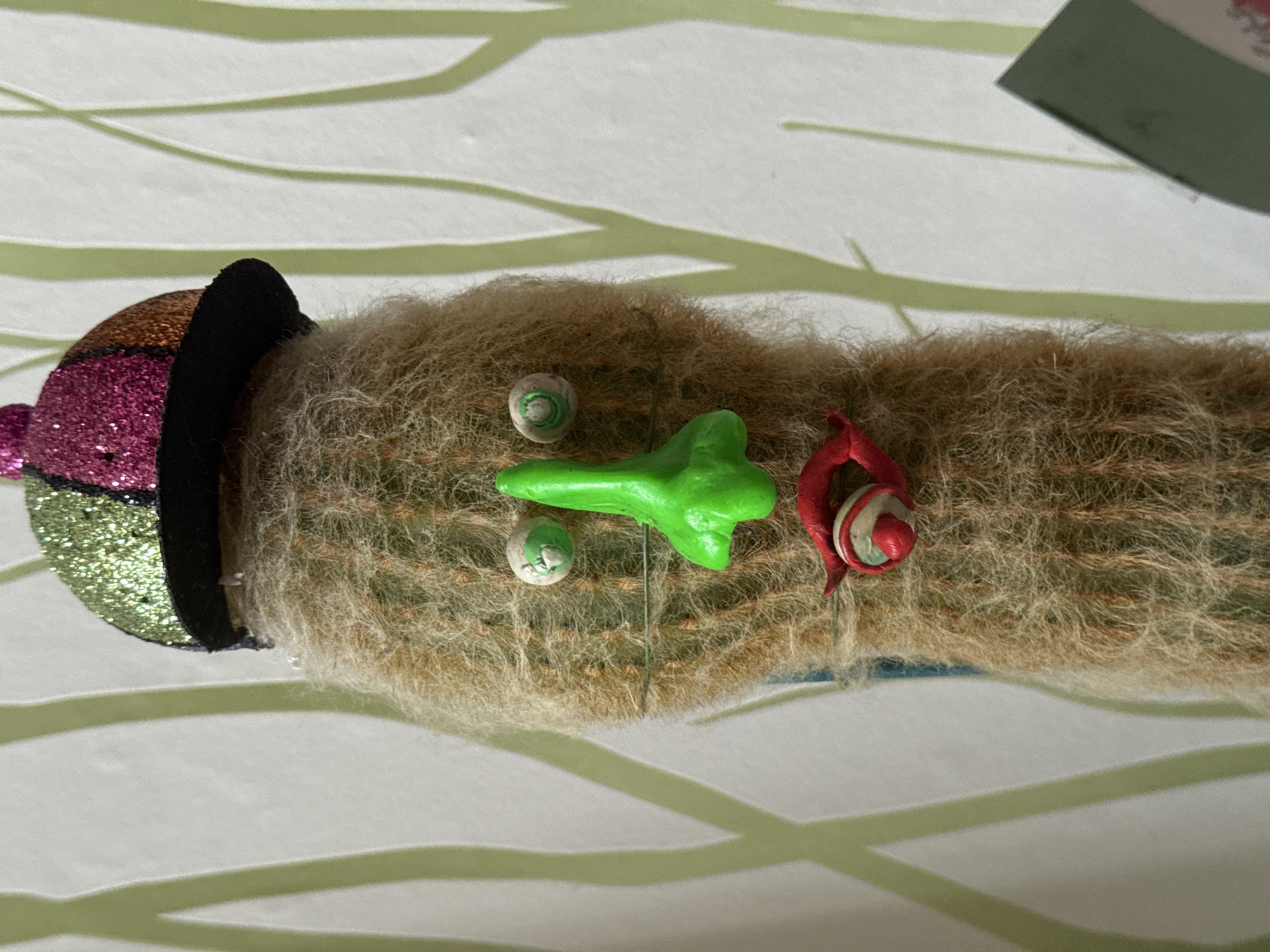 A close-up, still-life photograph features a long, cylindrical cactus covered in fluffy, tan spines. It is adorned with whimsical decorations: a sparkly, segmented hat in shades of green, pink, and bronze with a black brim sits at the left end of the cactus; a small, lime-green, blob-shaped object with a visible metal pin sticking out from the center rests on the cactus's surface; two small, spherical objects with green concentric circles are placed near the green shape; and a red, coiled decoration sits at the right end. The cactus is placed on a surface that appears to be light green with a white striped pattern. The image suggests a festive or decorative use of the cactus. There is no indication that the image was taken in Ново-Никольское, Russia.