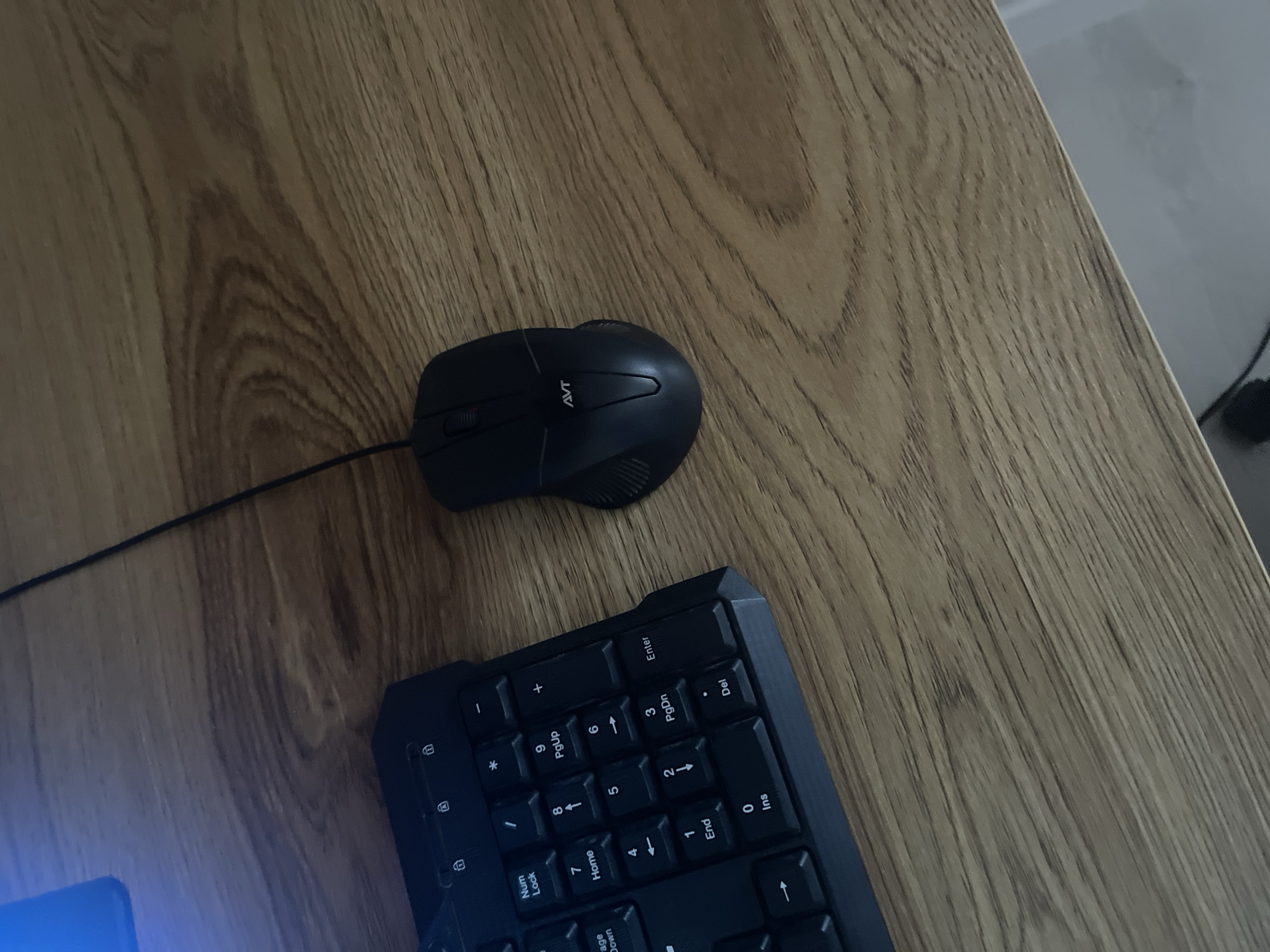The image displays a close-up, overhead view of a black computer mouse and a portion of a black keyboard resting on a wooden surface. The mouse is positioned to the right of the keyboard, with its cable extending upwards and to the left. The keyboard's numeric keypad is visible, showing keys labeled with numbers, arrows, and functions such as "Num Lock," "Home," "End," and "PgUp/PgDn." The wooden surface has a distinct grain pattern. No people are visible in the image. The scene appears to be a typical desk setup. There is no information within the image to suggest its location in Qarshi, Uzbekistan.