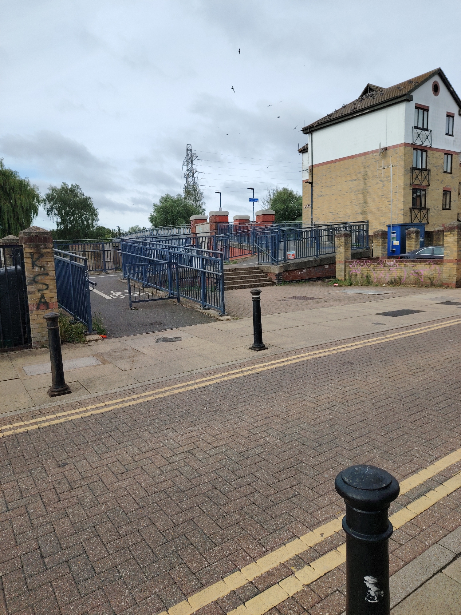 The image depicts an outdoor urban scene on an overcast day in Peterborough, United Kingdom, specifically near the Embankment Road / London Road area, providing access to the Riverside Walk along the River Nene.
In the immediate foreground, a textured brick road surface is visible, bordered by double yellow lines and a light grey paved sidewalk. Two black bollards are positioned on the sidewalk; one is prominent in the lower right, and another is further back on the left.
The midground features a blue-railed ramp and staircase structure, designed for pedestrian and cycle access, leading upwards towards a bridge or elevated pathway. A white wheelchair symbol is painted on the asphalt path of the ramp. To the left of the ramp, a brick pillar bears graffiti spelling "KCSA" in white letters, adjacent to a black metal gate. Another series of brick pillars and steps are visible on the right side of the ramp. A blue recycling bin and a dark-colored sedan are parked near the base of the residential building.
In the background, a multi-story residential building dominates the right side of the frame. It features a light yellow brick lower section and a white upper section, topped with a dark, gabled roof. Numerous birds are perched along the ridge and slopes of the roof. The building has multiple dark-framed windows, some with decorative black metal balconies. To the left of the building, a dense cluster of green trees is visible, with an electricity pylon and power lines extending upwards into the sky. The sky is largely overcast with light grey clouds, and several birds are seen flying at various altitudes.