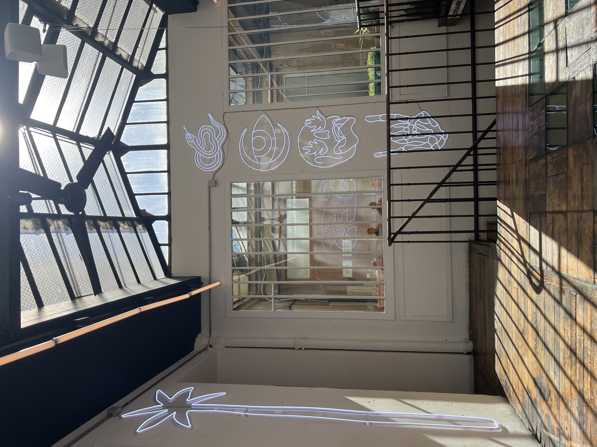 The image shows the interior of a building with a glass ceiling, likely an atrium or conservatory, in Paris, France. Natural light streams through the ceiling, creating dramatic shadows on the wooden floor. White neon lights in the shapes of a palm tree, a snake, an eye, a skull, and a hand making a rock-and-roll gesture adorn the white walls. A metal railing separates a walkway from a lower level. Through a window, a courtyard with potted plants and architectural details is visible. Several people are present, some appearing to be patrons or staff, though their faces are indistinct due to lighting and distance. The overall atmosphere suggests a contemporary, art-filled space.