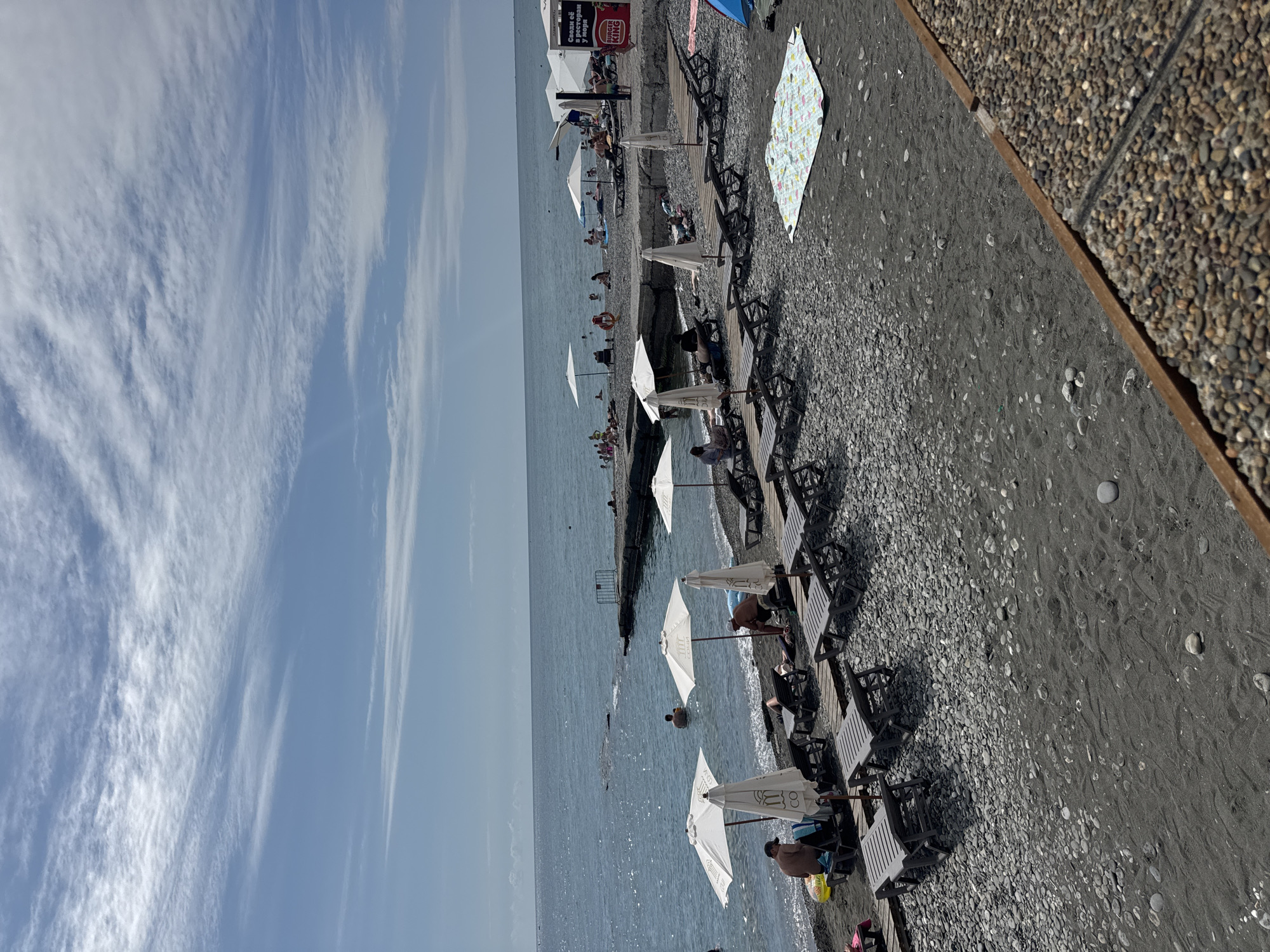 A wide-angle shot captures a bustling beach scene on a sunny day in Sochi, Russia. The foreground is dominated by a dark, pebbly and sandy shore, lined with rows of empty, dark grey lounge chairs and white beach umbrellas. A colorful patterned blanket is spread out on the sand to the right. Further back, people are scattered across the beach and in the calm, blue water. Several individuals are seated on lounge chairs under umbrellas, while others are either standing or wading in the sea. A concrete pier juts out into the water in the mid-ground, with a few people visible on it. The vast expanse of the Black Sea stretches to the horizon, meeting a clear blue sky dotted with wispy white clouds. A red and white Burger King sign is visible on a structure to the far right of the frame, indicating commercial activity near the beach. The overall impression is one of a popular, recreational destination.