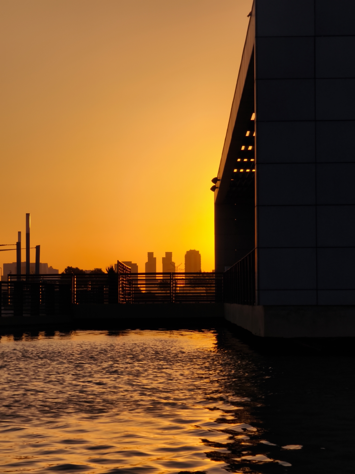 An outdoor, eye-level, vertical medium shot captures a sunset over a cityscape with a prominent modern building and a body of water in the foreground. The sky is a vibrant orange, transitioning to yellow near the horizon. Silhouetted against the sky are the outlines of various buildings and structures. In the foreground, a body of water reflects the warm hues of the sunset, creating a rippling pattern of light and shadow. To the right, a large, dark, modern building with clean lines and rectangular panels occupies a significant portion of the frame, its geometric form contrasting with the softer lines of the cityscape and the natural elements. The building is partially illuminated by lights on its underside, adding to the dramatic effect. A dark railing runs along the edge of the building's elevated platform. The scene conveys a sense of urban tranquility and the beauty of natural light in an architectural setting. The location is the rooftop of the Soroka Medical Center in Be'er Sheva, Israel, specifically overlooking the western part of the city as the sun sets.