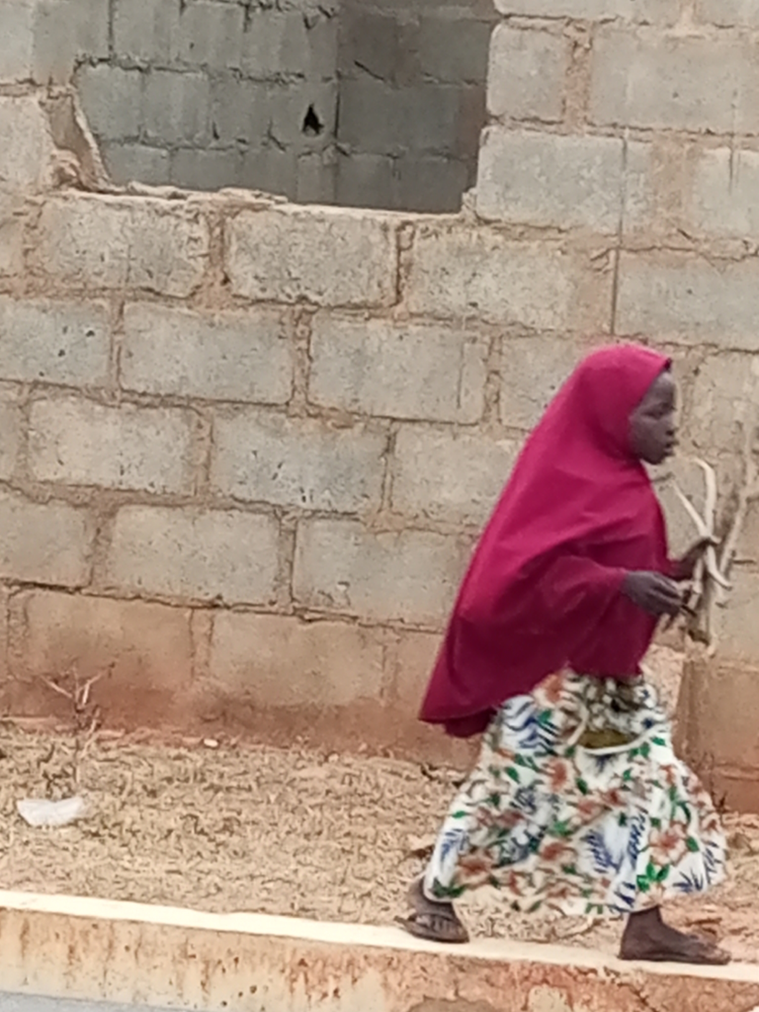 A child walks across a dirt and gravel area in front of a brick building. The child wears a red hijab and a white skirt with a floral pattern. The child is carrying a bundle of sticks in their right hand. The brick building appears to be under construction, with unfinished sections and a hole in the wall. The ground is covered in loose dirt, gravel, and some scattered debris.