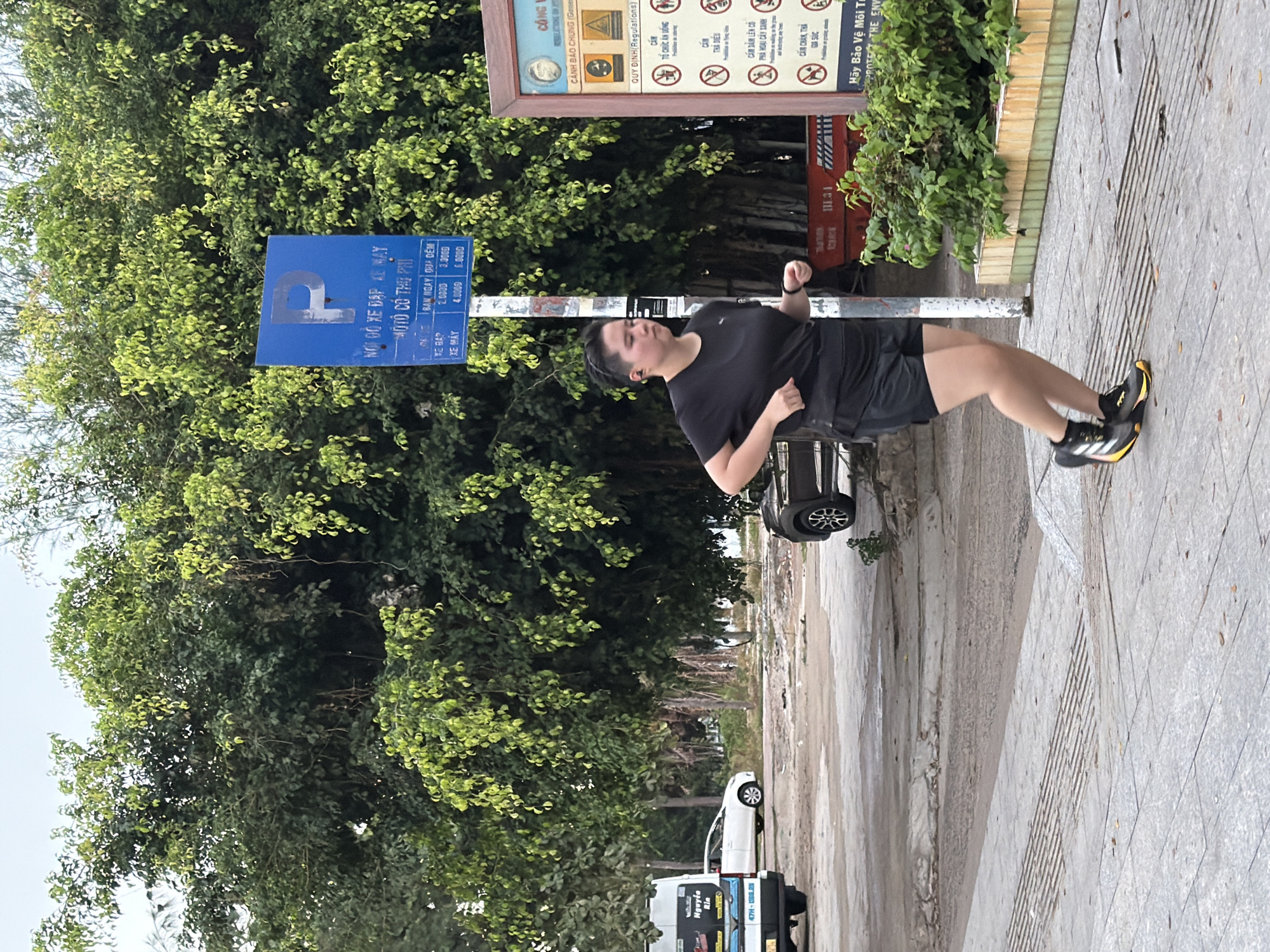 The image shows a person jogging on a paved walkway in Nha Trang, Vietnam. The person is wearing a black t-shirt and black shorts, with black and yellow athletic shoes. Behind the jogger, there is a large tree with dense green foliage. To the left of the tree, a blue parking sign with white text is visible. Further to the left, a white van with text and graphics on its side is parked. In the background, there is a white car. To the right of the jogger, a sign with text and icons is mounted on a pole, next to a planter with green plants. The ground is paved with light-colored square tiles. The sky is overcast.