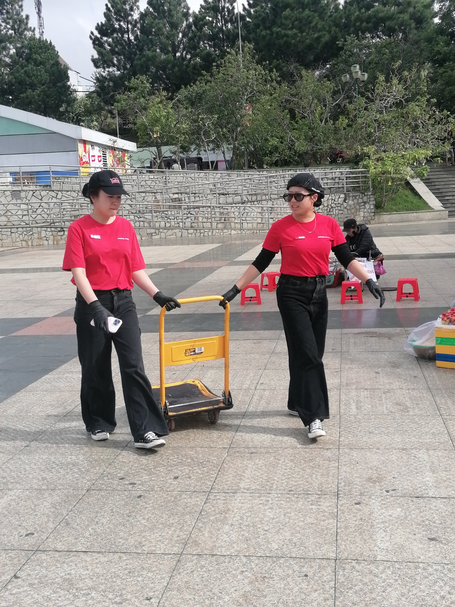Two young women wearing red t-shirts, black pants, black baseball caps, and black gloves are walking on a paved plaza in Da Lat, Vietnam. The woman on the left is holding a white smartphone in her gloved left hand, while the woman on the right is pushing a yellow trolley with her gloved hands. The trolley has a black base and yellow handles. In the background, there are trees, a stone wall, a building with a light-colored facade, a street lamp, and red stools. There is also a person in a black jacket in the background. The ground is paved with light-colored square tiles.