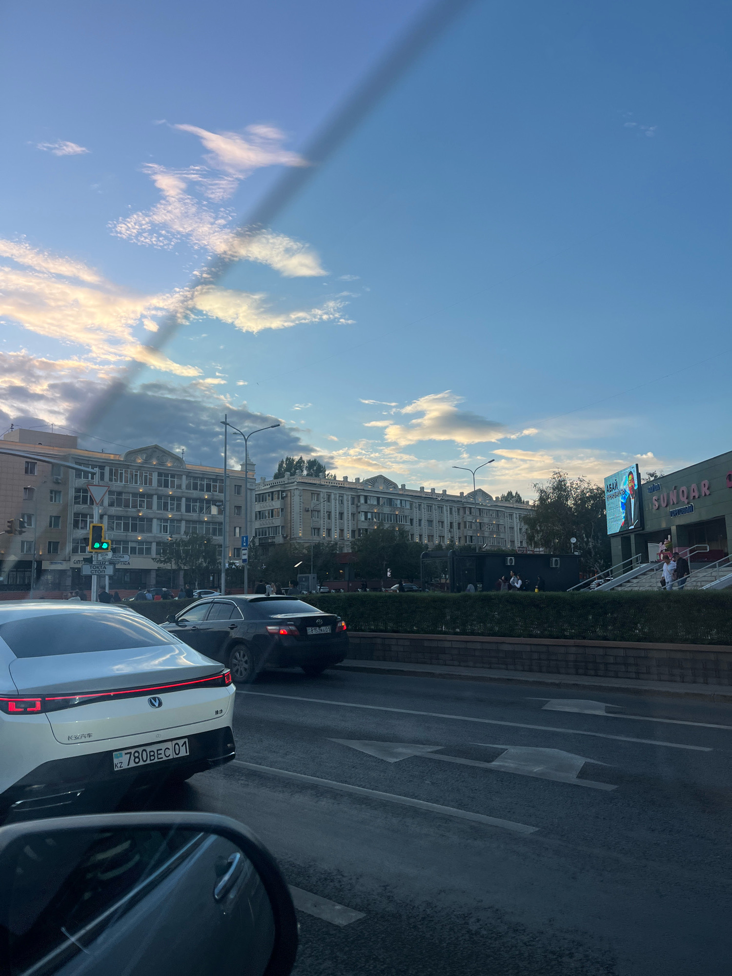 The image captures a street scene in Astana, Kazakhstan, likely during the late afternoon or early evening, given the sky's lighting. A white electric vehicle, with a distinctive rear light bar and the license plate "KZ 780BEC01," is in the foreground, facing away from the camera. To its right, a dark-colored sedan with license plate "815ZMA01" is also on the road.
The street is a multi-lane road with clear lane markings, suggesting it is a significant thoroughfare. Traffic lights are visible, with one displaying a green light.
Several buildings line the street. On the left, a light-colored multi-story building with numerous windows stands. In the center, a larger, more ornate building with architectural details is prominent. To the right, a building with a large digital screen displaying a portrait and text in what appears to be Kazakh language is visible. The screen reads "SUNQAR" in large letters.
People are seen walking on sidewalks and near the buildings, and some are gathered in small groups. The environment includes trees and other greenery bordering the sidewalk, creating a pleasant urban landscape. The sky is a mixture of blue with scattered white and gray clouds, with a visible shadow or streak across the upper portion of the image.