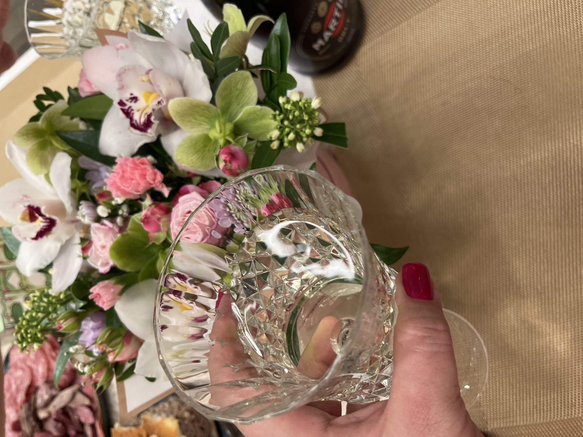 The image shows a hand holding a crystal glass in front of a floral arrangement. The arrangement includes orchids, carnations, and greenery. A bottle of Martini vermouth is visible in the background. To the left, there's a charcuterie board with various meats and cheeses. The background is a textured beige surface.