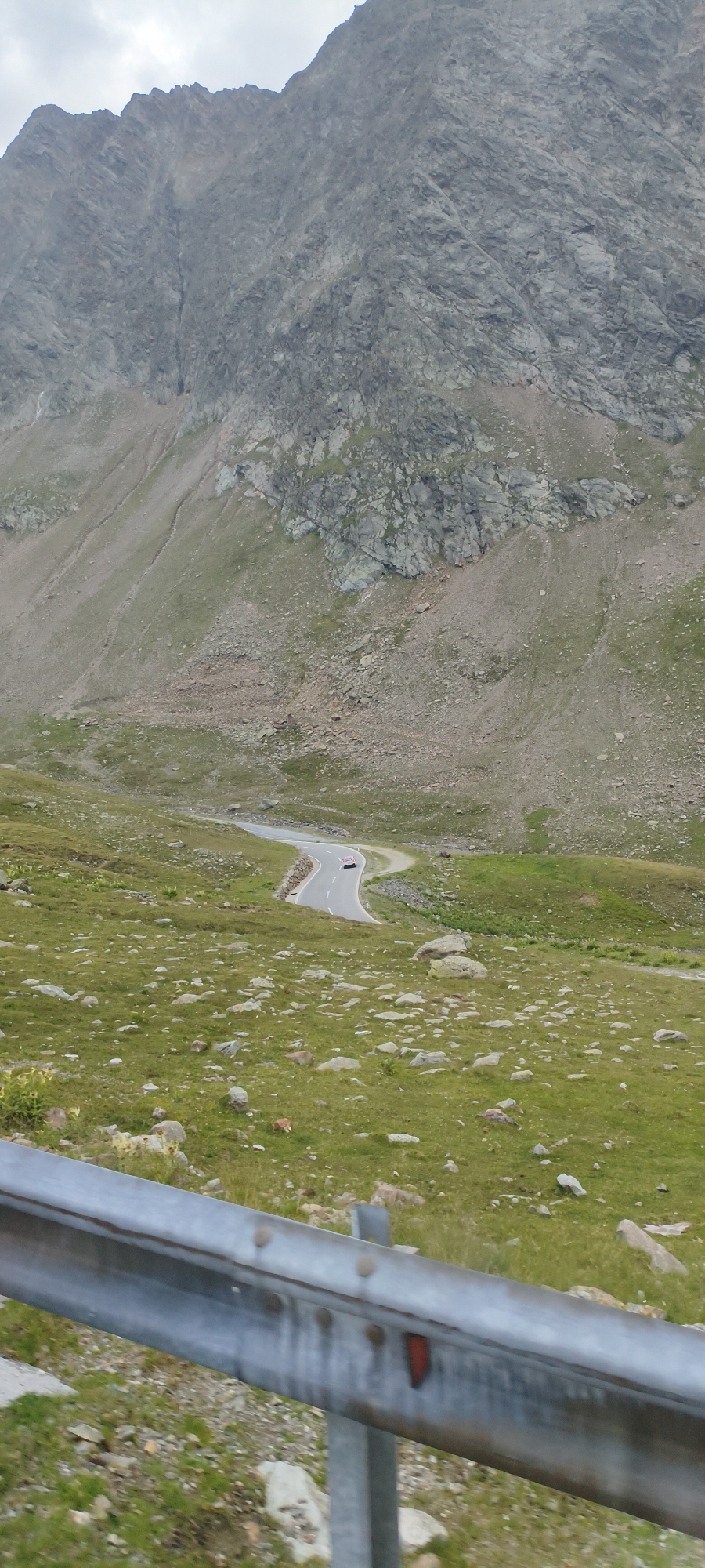 The image depicts a winding mountain road in Sölden, Austria, with a red and white car navigating a curve. The road is paved and bordered by a metal guardrail in the foreground. The surrounding environment is a rocky, grassy mountainside with sparse vegetation and scattered boulders. In the background, a large, steep mountain face dominates the scene under a cloudy sky. The image is captured from a slightly elevated perspective.