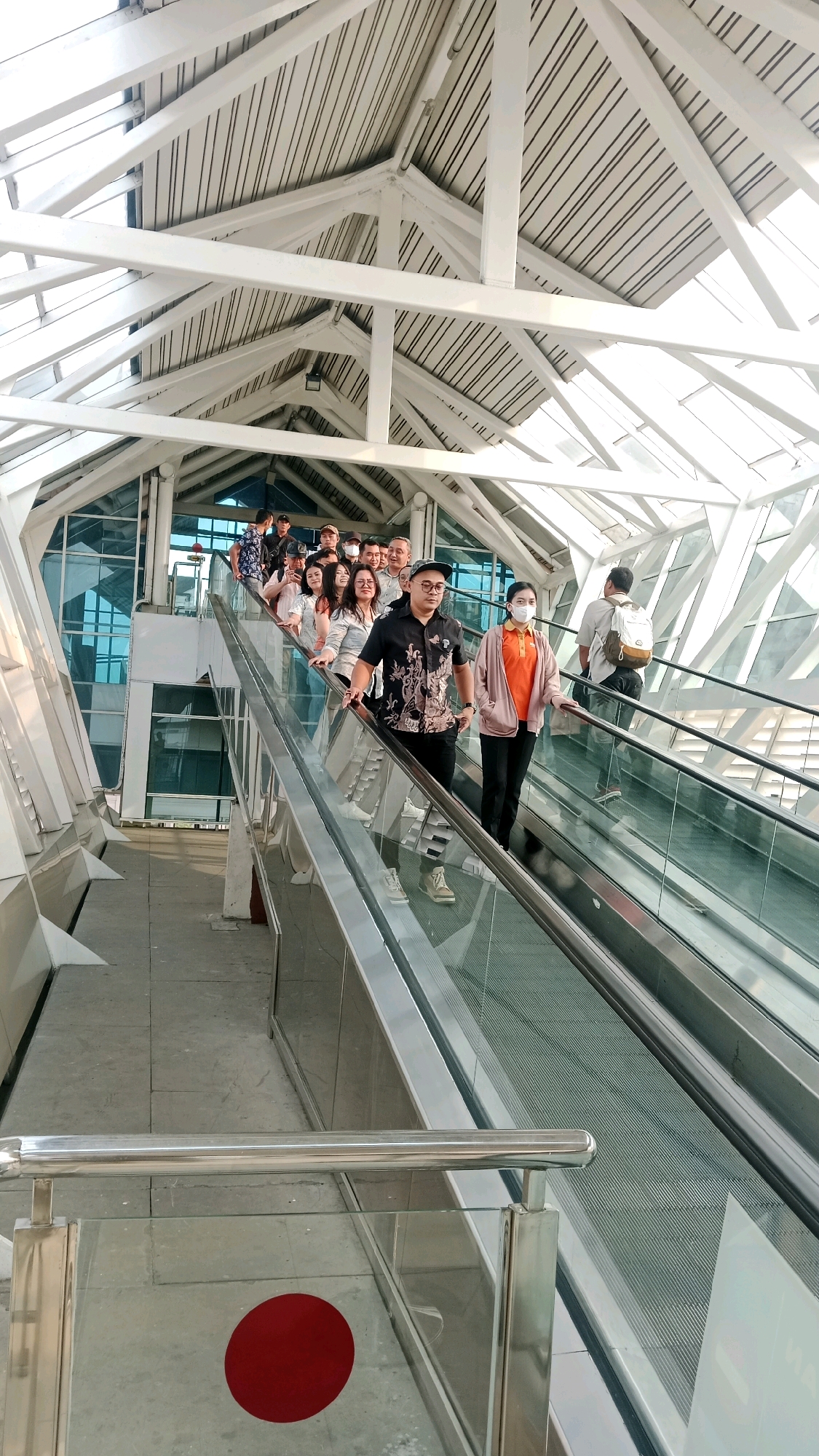 The image shows an escalator with several people on it. The escalator is enclosed by glass and metal railings. The ceiling is made of glass and white beams, allowing natural light to filter in. The people are dressed in casual clothing, and some are wearing face masks. In the foreground, a red circular marker is visible on the floor. The environment appears to be a modern public space, possibly an airport or a shopping mall. This location is identified as the Tangerang City 3D Trick Art Museum in Tangerang, Indonesia.