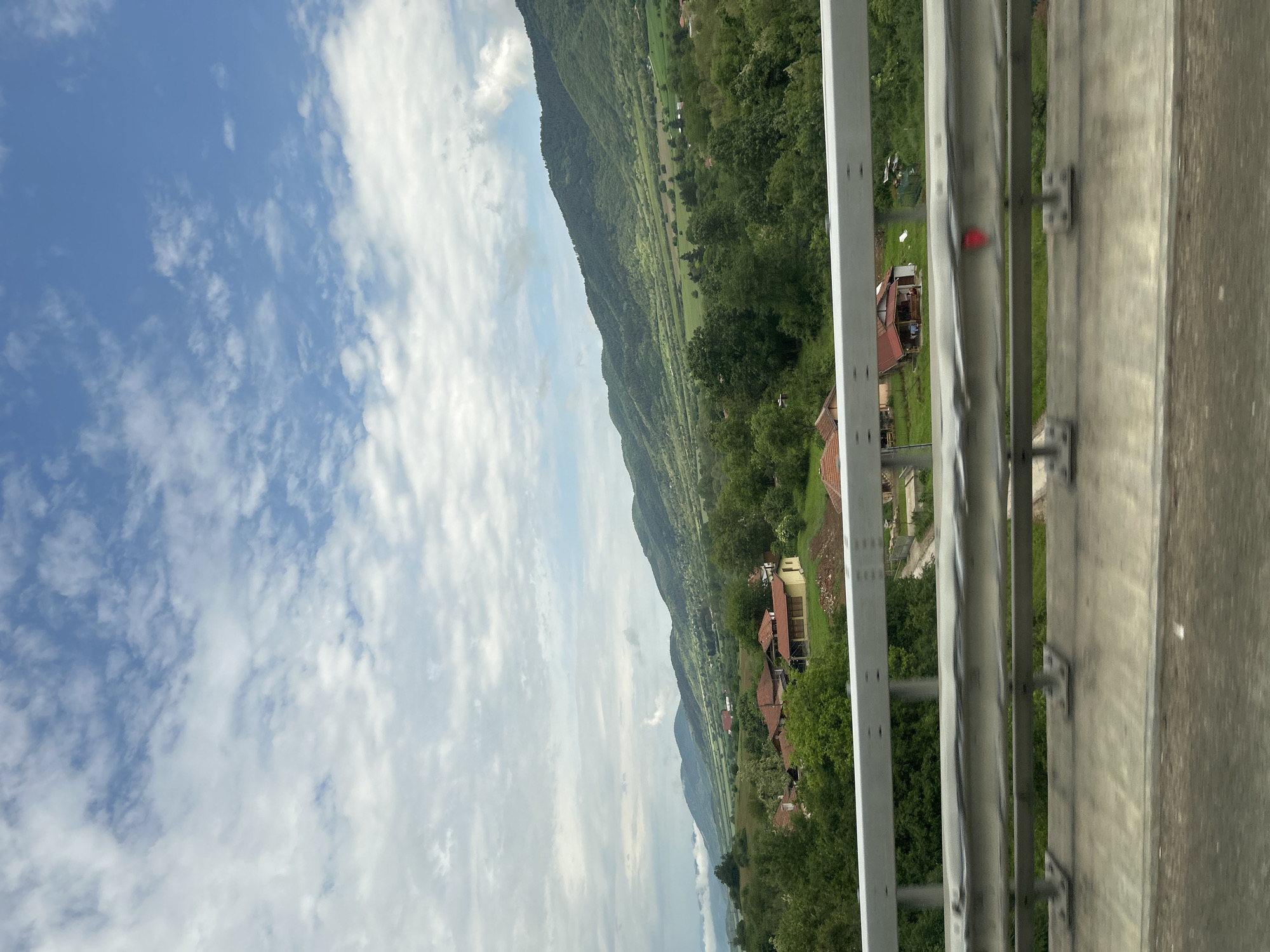 The image shows a view from a vehicle, likely a car, crossing a bridge or overpass. A metal guardrail is prominently in the foreground, running horizontally across the right side of the frame. The guardrail obscures some of the background but reveals a landscape of green fields, trees, and houses with red roofs. In the distance, there's a large mountain range under a sky filled with white, puffy clouds. The angle of the shot suggests the image was taken while moving, and the focus is on the scenery beyond the guardrail.