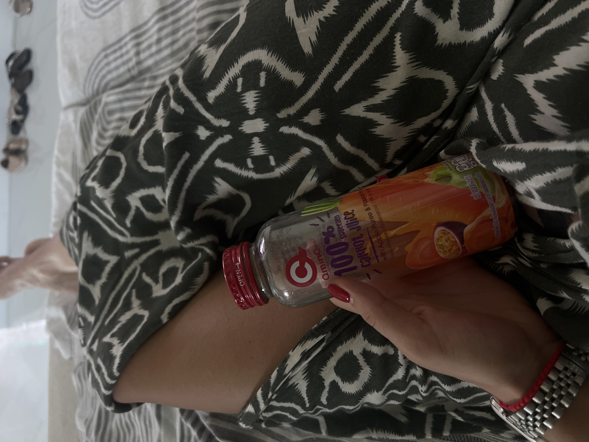 A close-up, eye-level shot shows a person's hand holding a bottle of "Q Amina 100% squeezed carrot juice with Apple Puree & Passion Fruit". The bottle has a red cap and a label with images of carrots and passion fruit. The hand has red nail polish and is wearing a silver watch with a red string bracelet. The person is lying down on a bed with grey and white striped sheets and a dark green and white patterned throw blanket. The background is blurred, but shows a few pairs of shoes on the floor, suggesting the setting is indoors, possibly a bedroom in a residential area of Choeng Thale, Thailand. The lighting appears to be natural, coming from a window or an open door.