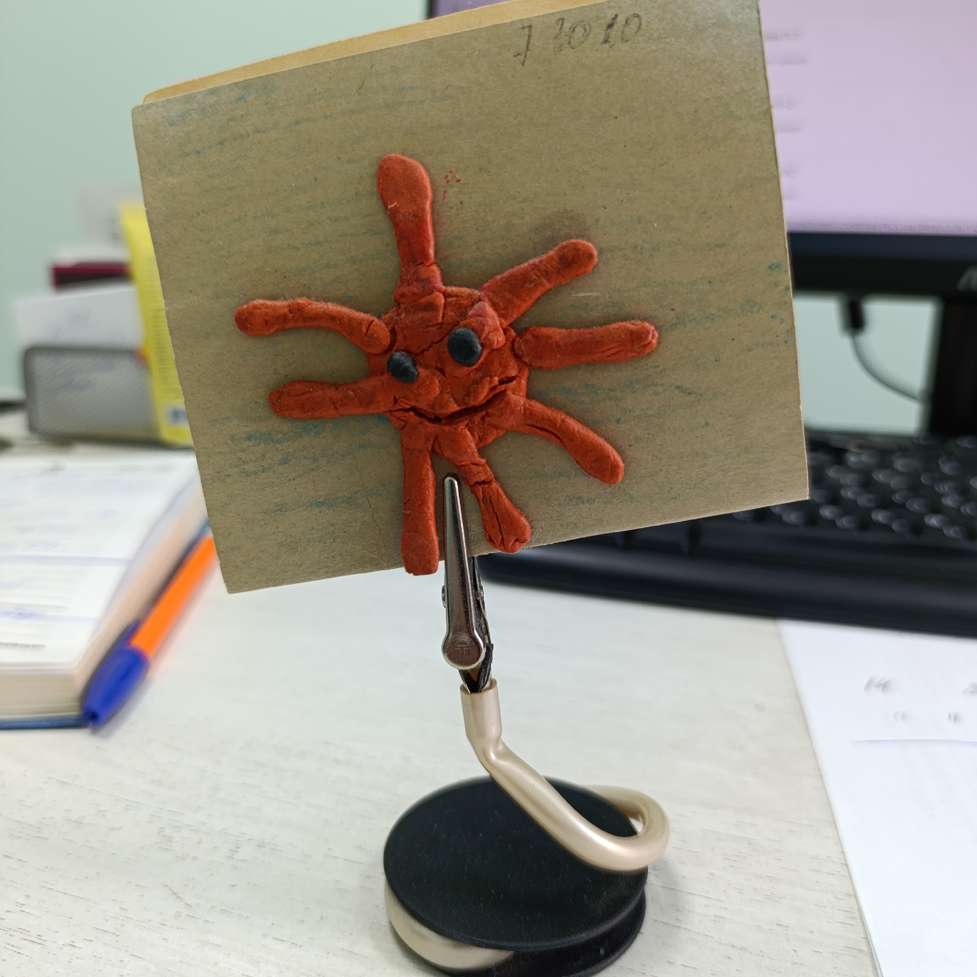 The image depicts an office desk setup in Марковский, Russia. A handcrafted artwork is prominently displayed in the foreground, consisting of an orange clay figure affixed to a rectangular piece of brown cardstock. This cardstock is held vertically by a silver crocodile clip attached to a flexible, light beige, curved arm, which in turn is mounted on a circular black base.
The orange clay figure has an irregular, roughly star-shaped or amoeboid form with approximately seven distinct appendages radiating from a central body. Two small, spherical black elements are embedded in the upper part of the central body, resembling eyes. The clay surface exhibits texture, minor cracks, and irregularities. A small red smudge is visible on the cardstock directly above the clay figure.
The brown cardstock, appearing to be cardboard or heavy paper, features faint blue ink marks or lines across its surface. In the upper right corner, the handwritten inscription "7.10.10" is legible in dark ink.
The desk surface beneath the object is light-colored, possibly white or light grey, with a fine grain texture. In the blurred background to the left, a blue and orange pen rests on a stack of white papers or a book, next to a yellow book or folder. To the right, a black computer keyboard is partially visible, behind which a monitor displays a blurred, light-colored screen with indistinct text. A white sheet of paper is also present on the desk to the right of the stand's base. The scene is illuminated by what appears to be uniform indoor artificial lighting.