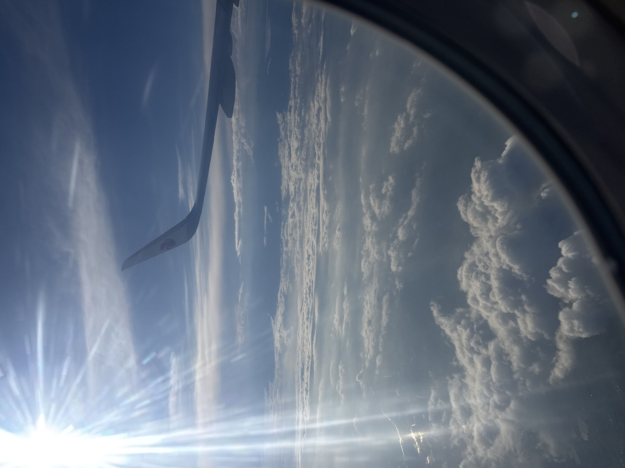 The image shows a view from an airplane window. The sky is visible, with some clouds. Part of the airplane wing is also visible in the frame. The sun is shining brightly, causing a glare. The location is not discernible as Tashkent, Uzbekistan.