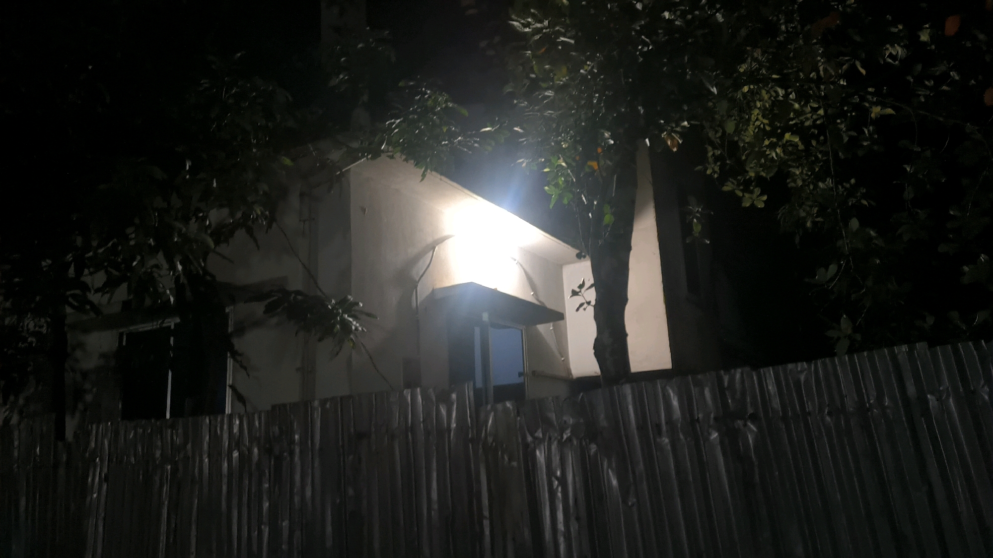 A nighttime exterior shot shows a portion of a building in Begumganj Upazila, Bangladesh. A bright light illuminates the facade of the building, which appears to be made of concrete. A dark-colored door is visible beneath an overhang. Trees with dark leaves are situated in front of and to the sides of the building. A corrugated metal fence runs across the foreground. The scene is dark, with the exception of the illuminated building.