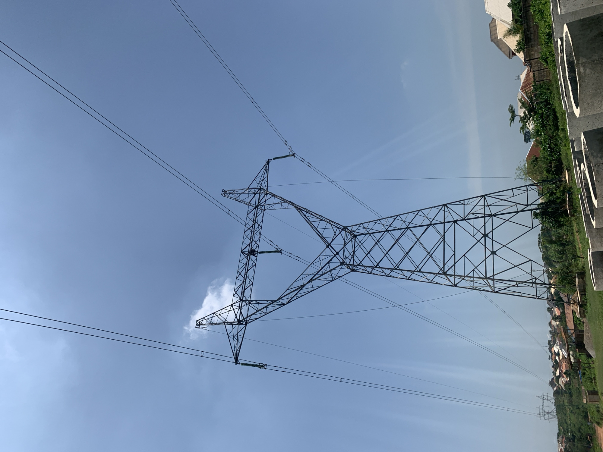 A tall, black metal electricity pylon stands prominently in the foreground, its lattice structure silhouetted against a clear blue sky. Multiple high-voltage power lines extend from the top of the pylon, stretching across the frame. In the background, a dense residential area with houses featuring red and white roofs is visible, nestled on a grassy, undulating landscape. Some construction materials, possibly concrete rings, are scattered in the lower right foreground. The scene appears to be outdoors on a sunny day, with the sky showcasing a few wispy clouds and sun rays.