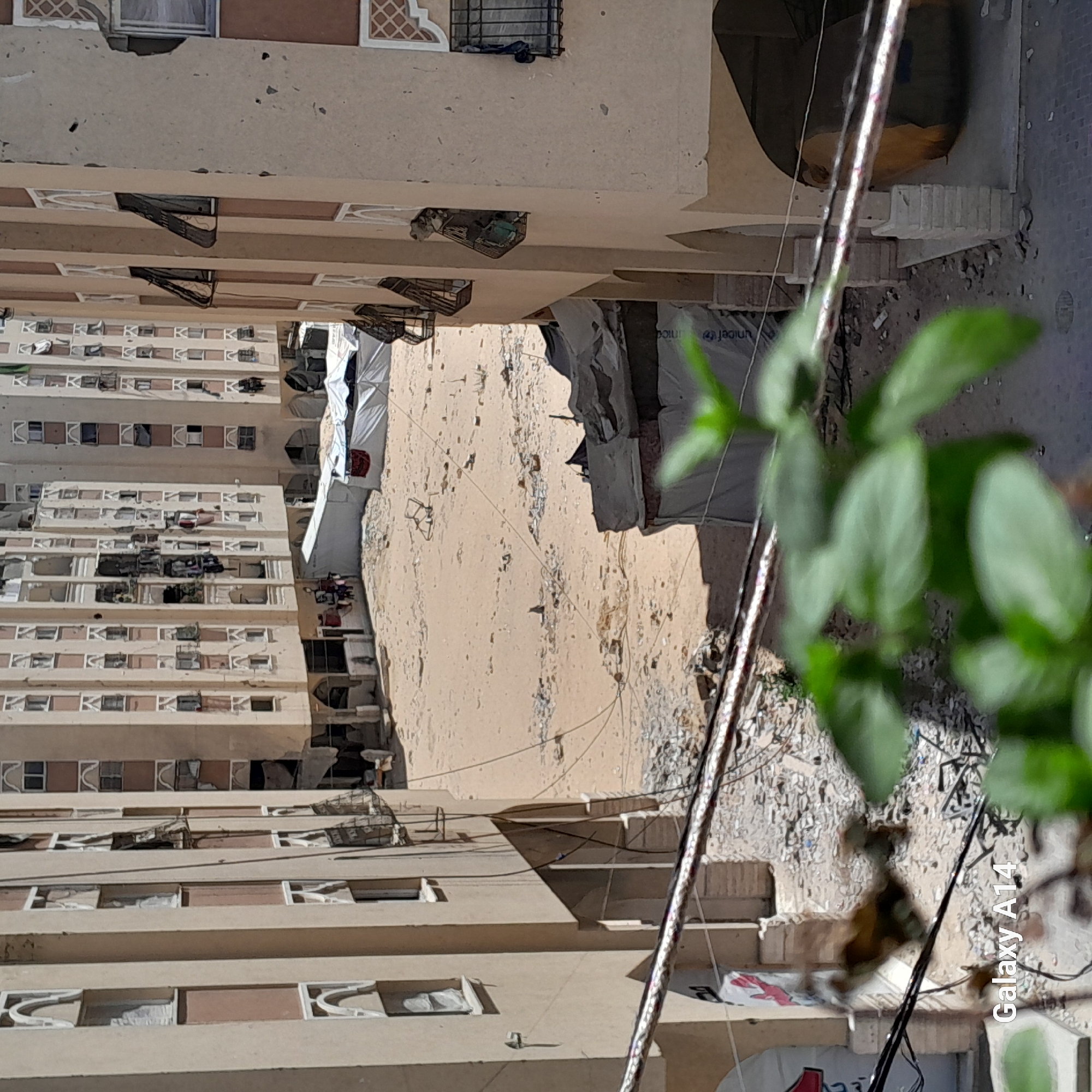 The image shows a view of damaged apartment buildings in Khan Yunis, Palestinian Territories. The buildings appear to be multi-story residential structures with balconies and numerous windows. Some windows are broken or have debris around them, and parts of the buildings show signs of structural damage, with visible cracks and missing sections of facade.
In the foreground, a rope or cable stretches horizontally across the frame, partially obscuring the view. A blurry green plant is also visible in the lower right corner, suggesting the photo was taken from a window or balcony.
Between the buildings, there is an open area covered in sand and debris. Several white tents or temporary structures are set up in this area, indicating potential displacement or temporary shelter. There are also scattered bricks and rubble, further suggesting damage. Electrical wires are visible, running between the buildings and across the open space.
The overall scene depicts an environment affected by conflict or destruction, with a focus on the urban landscape of residential buildings and the surrounding area.